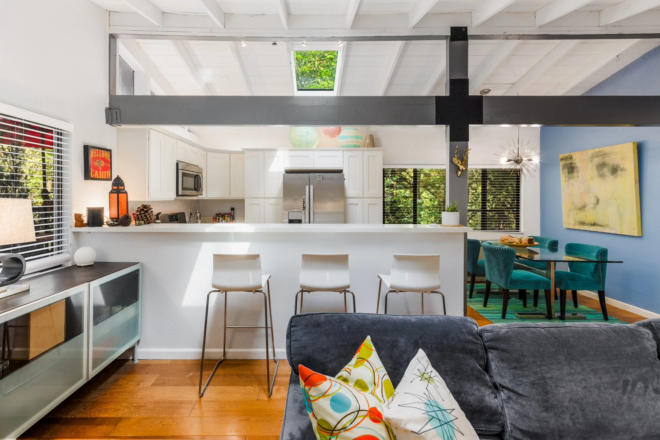 Open-concept kitchen and dining area with white cabinets, stainless steel refrigerator, colorful pendant lights, and a breakfast bar with three white chairs. Blue accent wall with artwork, houseplants, and a dining table with teal chairs. Large skyli