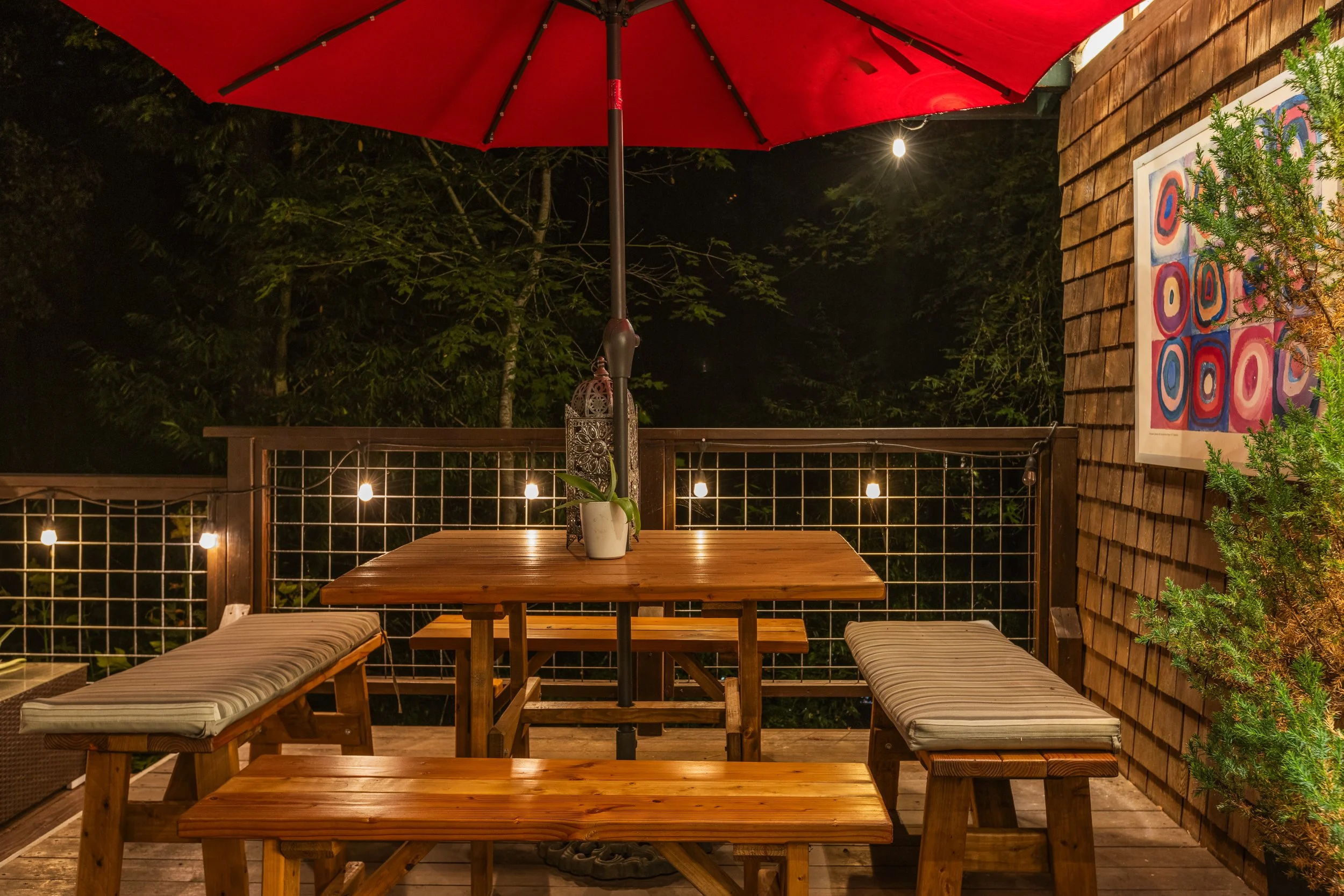 A cozy outdoor patio at night featuring a wooden table with two benches, a red patio umbrella, string lights, a potted plant, and a colorful abstract painting on a wooden shingle wall, with trees and a single bright light in the background.