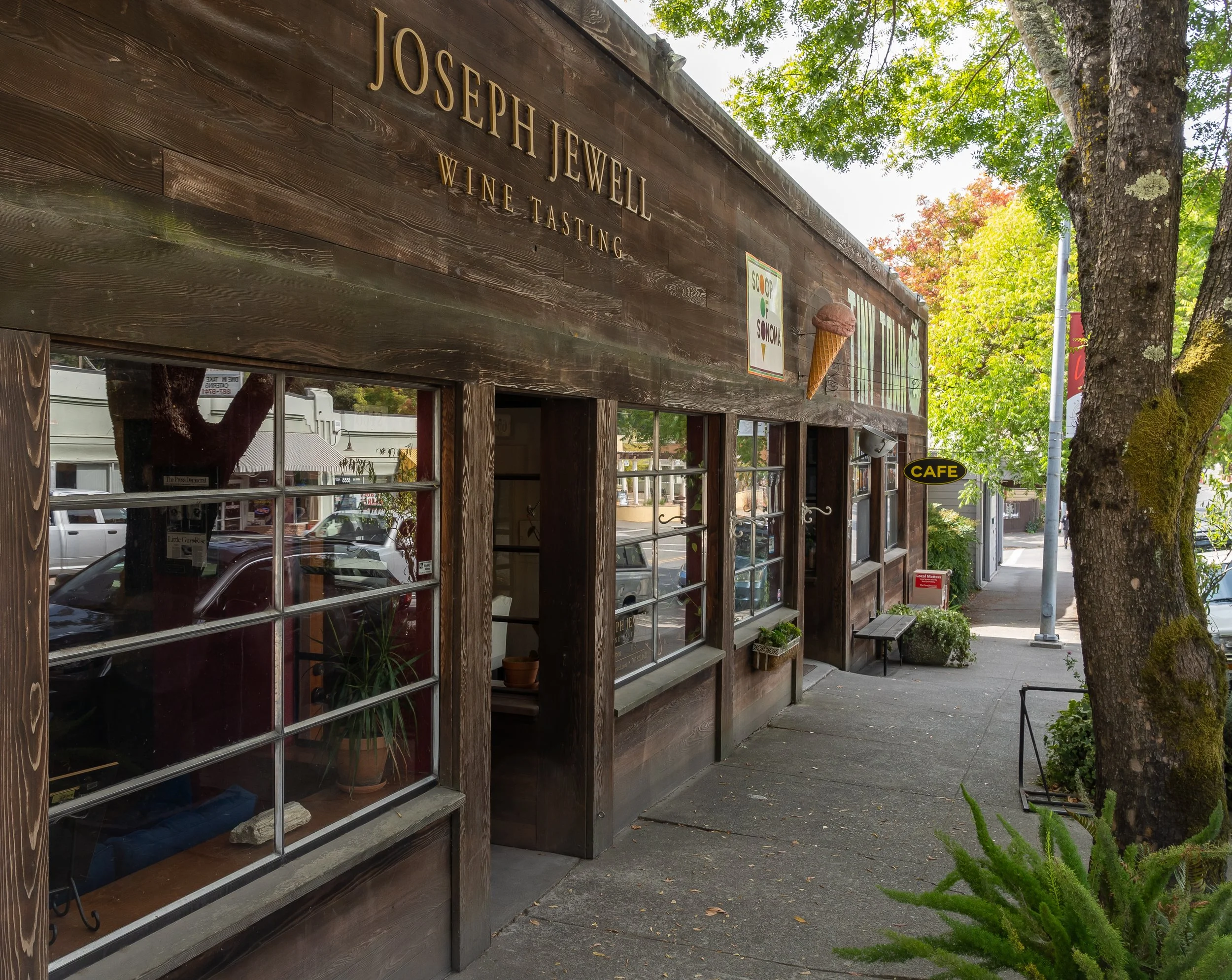Exterior of a wooden building with gold letters reading "Joseph Jewel Wine Tasting" and an ice cream cone sign. Reflective window showing parked cars and street lights, surrounded by trees with green foliage.