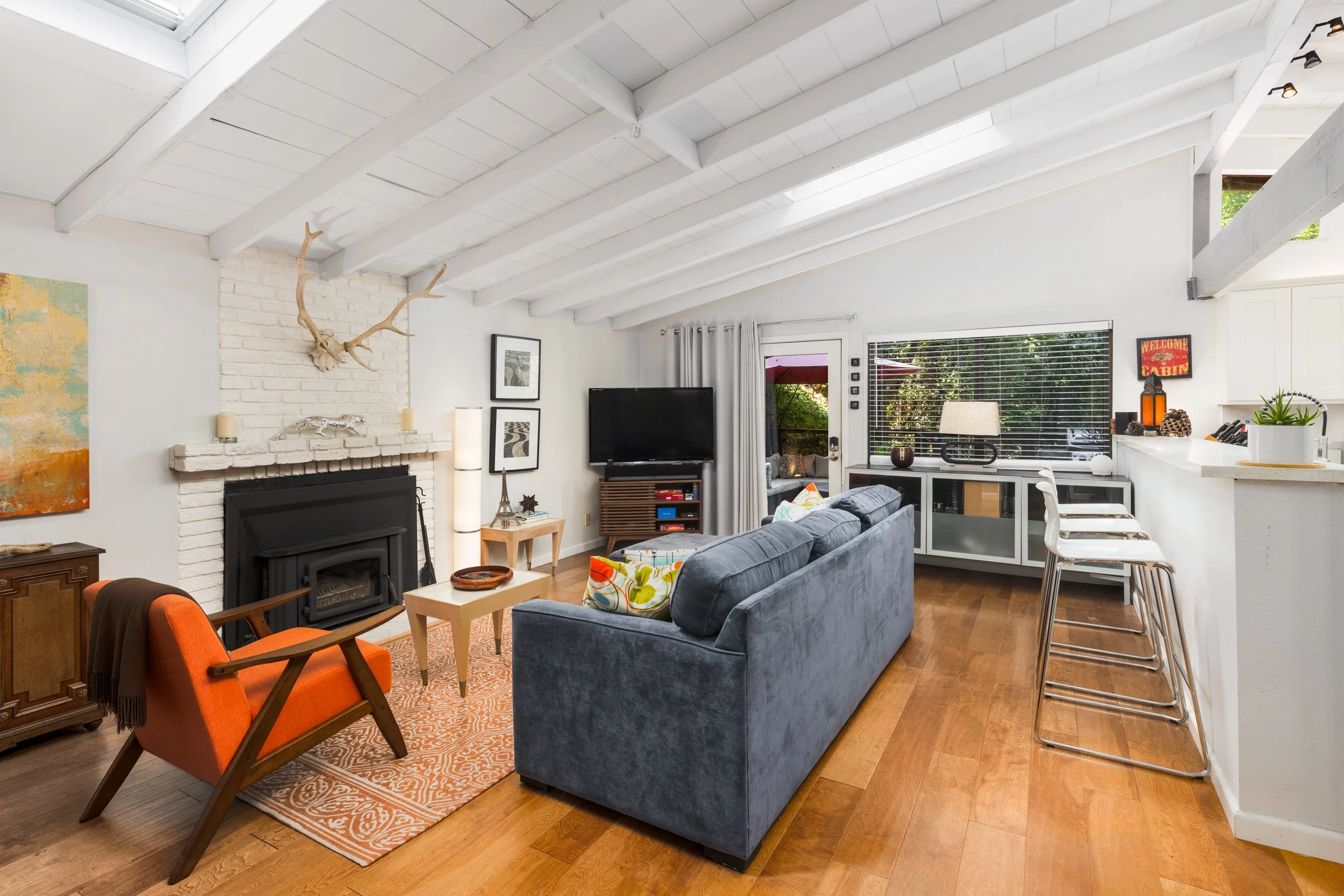 Modern living room with white vaulted ceiling, white brick fireplace, mounted antlers, and a flat-screen TV. Features a blue sofa, orange armchair, patterned rug, and a kitchen counter with bar stools. Large windows with blinds and curtains reveal gr