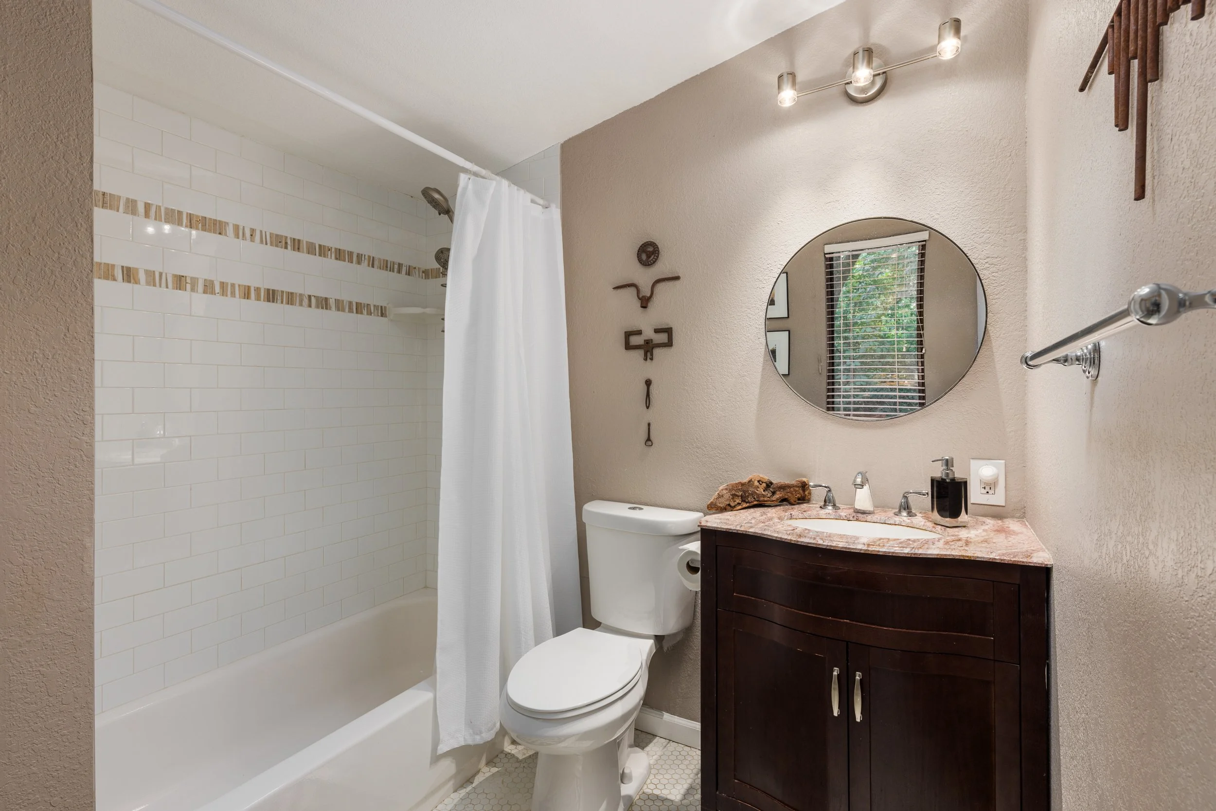 Bathroom with a shower, toilet, and vanity with a round mirror, window with blinds, and wall decorations.