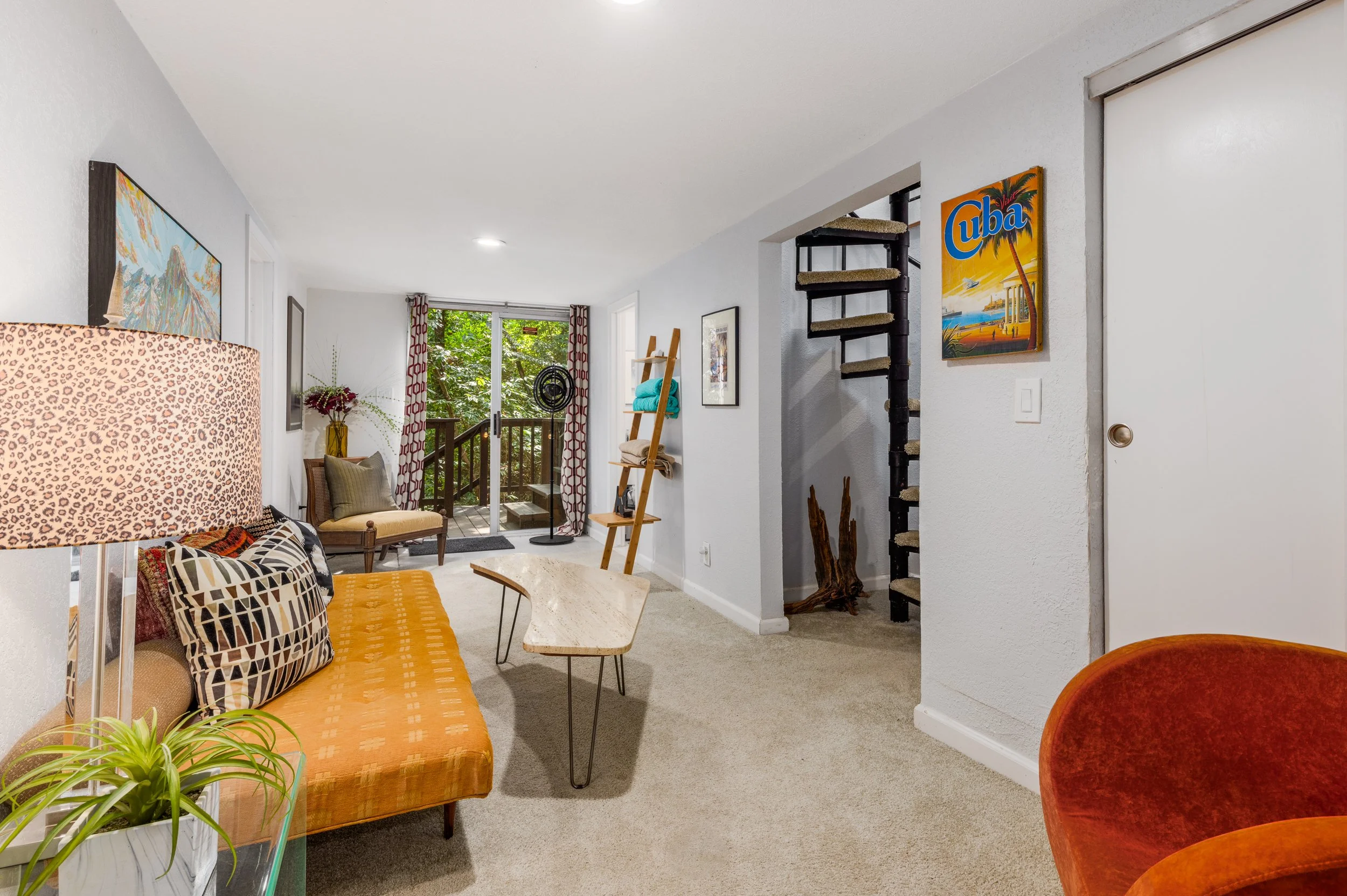 Cozy living room with colorful patterned pillows, orange sofa, a beige armchair near a sliding glass door, plants, and artwork on white walls.