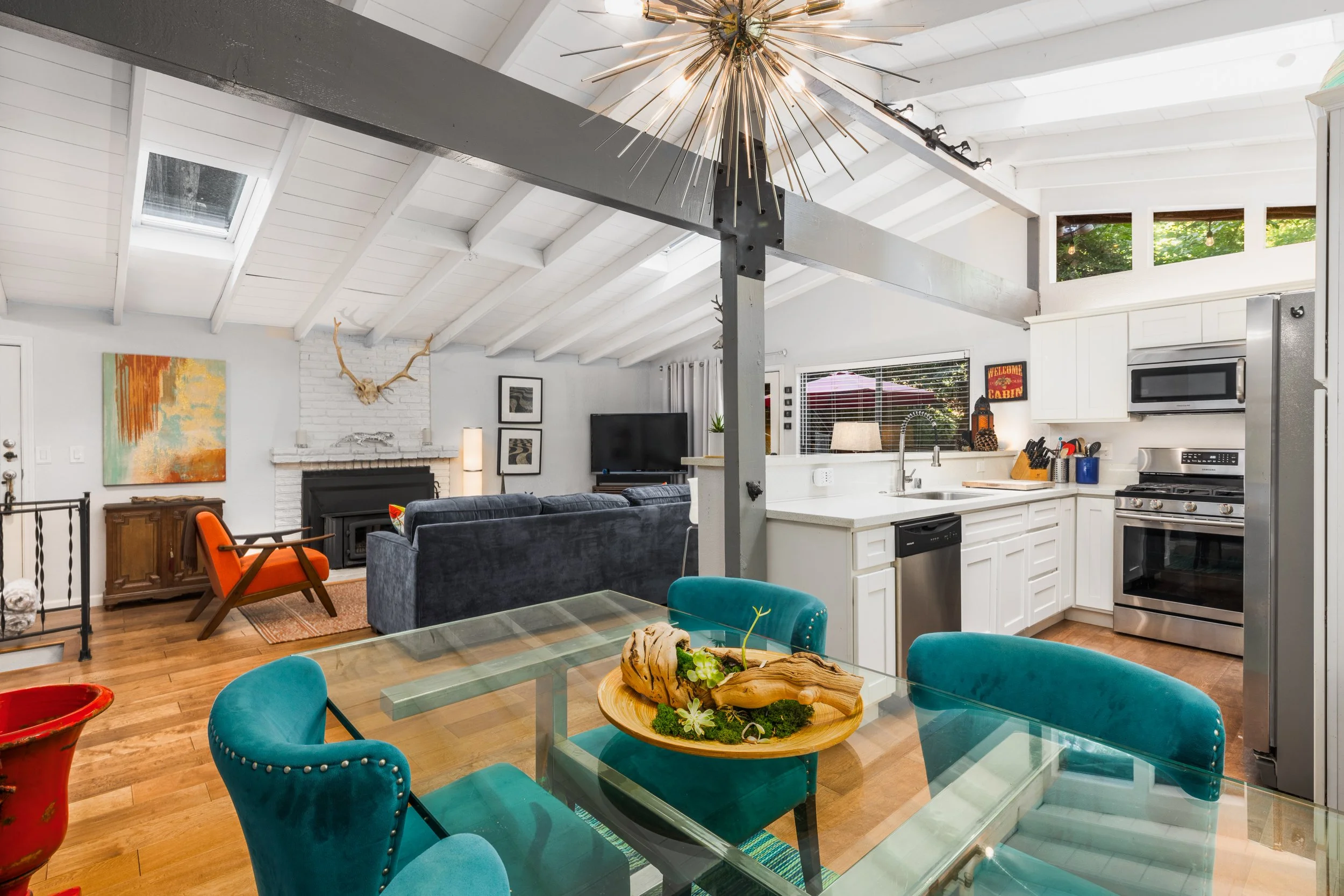 Interior view of a modern, open-plan kitchen and living room with high, white, vaulted ceilings. The kitchen features white cabinets, stainless steel appliances, and a large window with trees outside. The living area has a fireplace, a television, an
