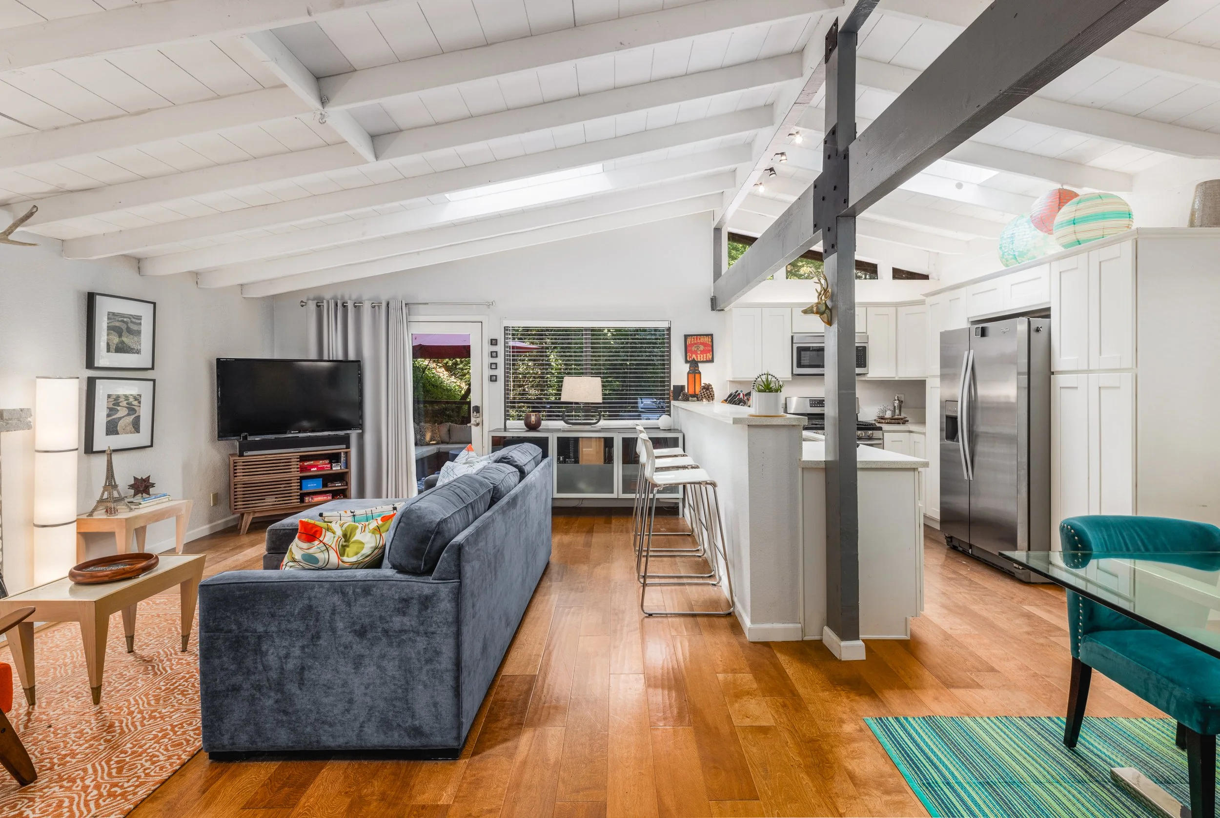 Interior of a living space with white vaulted ceiling, exposed wooden beams, a gray sofa with colorful pillows, a small wooden side table, a flat-screen TV, a window with blinds, and a kitchen area with white cabinets and stainless steel appliances, 
