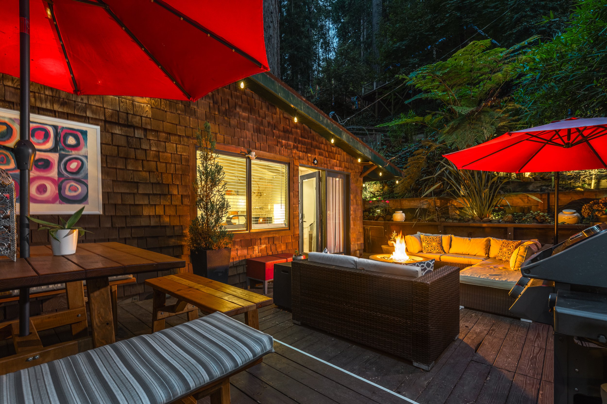Outdoor backyard patio area at dusk with wooden decking, string lights, patio furniture with cushions, a fire pit, large umbrellas, potted plants, and a view of trees in the background