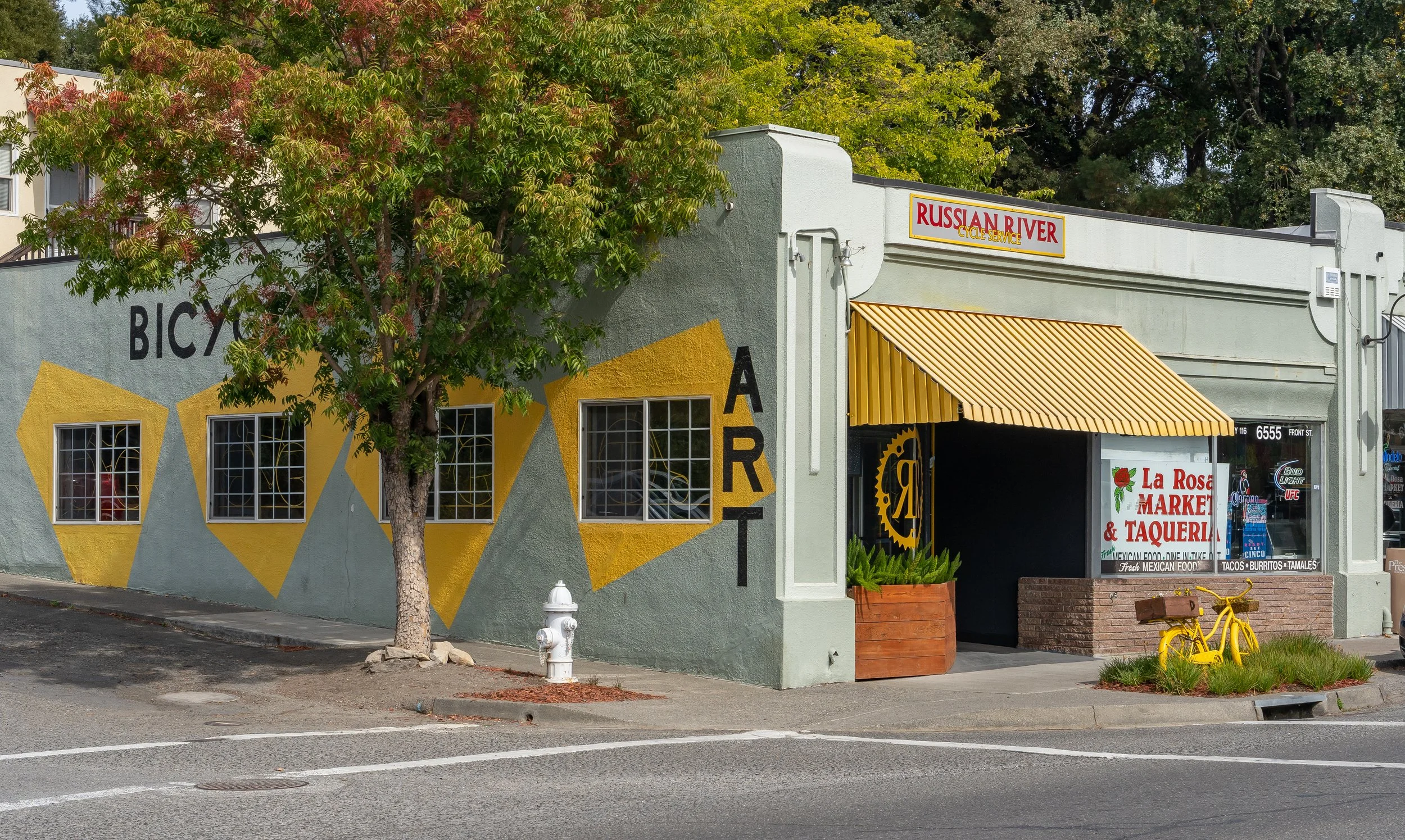 Street view of a building with a yellow awning, a sign for Russian River Cycle Service, graffiti-style art on the wall, and a tree partially covering the building. There is a yellow bicycle and a sign for La Rosa Market & Taqueria in front of the bui