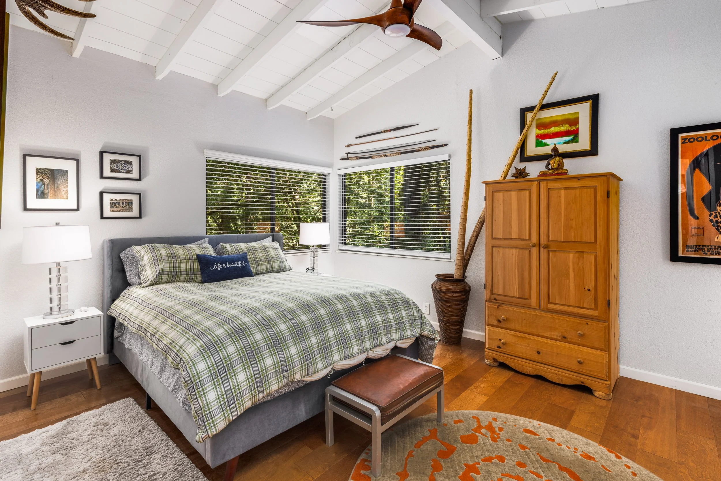 A cozy bedroom featuring a gray upholstered bed with plaid bedding and decorative pillows, white walls with framed artwork, a white nightstand with a lamp, a wooden chest at the foot of the bed, a wooden wardrobe, and a window with blinds showing gre