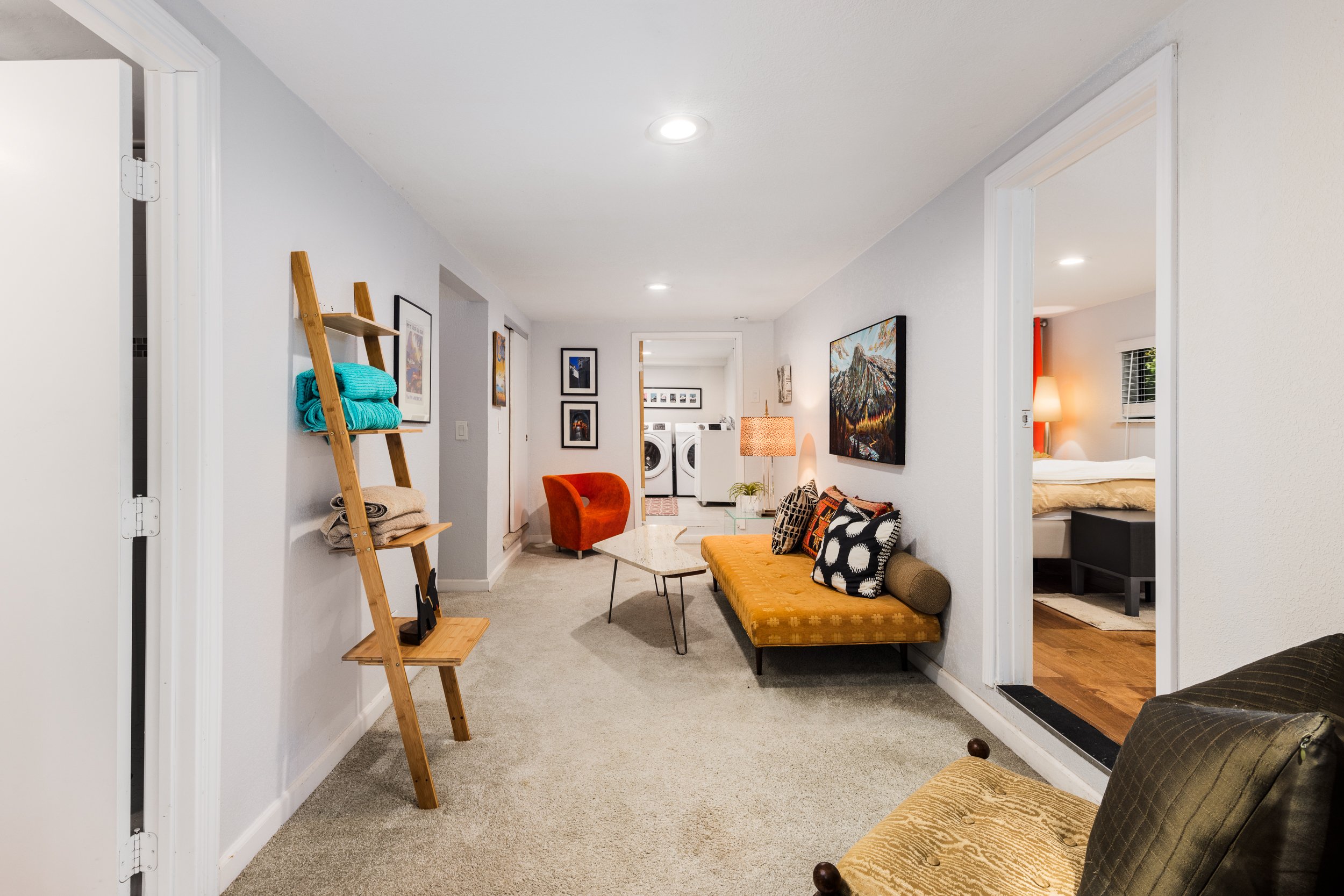 Living room with beige carpet, orange velvet couch, black and white pillows, TV, gallery wall, white washer and dryer in the background, and doorway leading to a bedroom with bed and window.