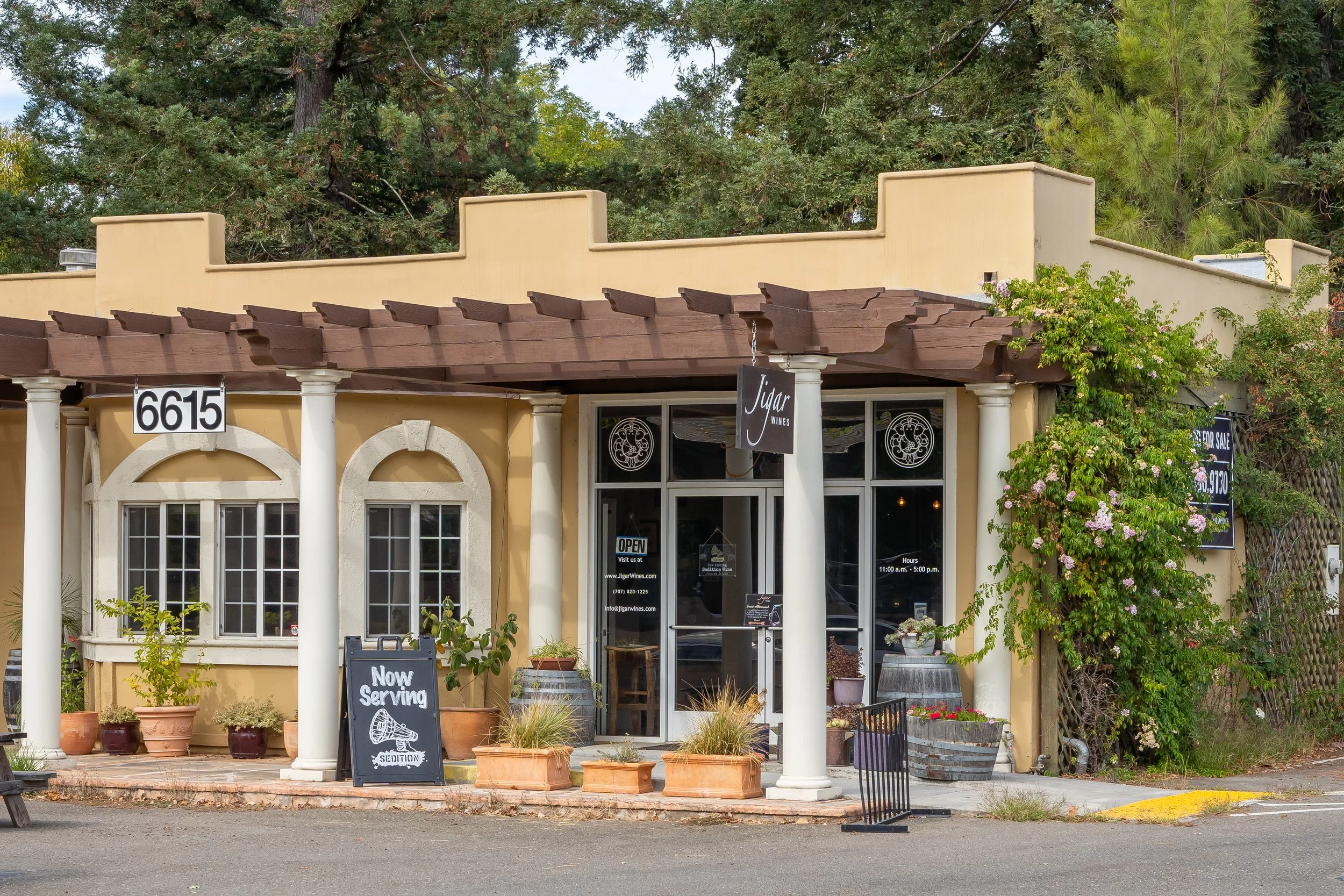 Front of a small restaurant with potted plants, a sign reading 'Now Serving', multiple windows, a door, and a blackboard sign. The building has a yellow exterior with white columns supporting an overhang. Trees are visible in the background.