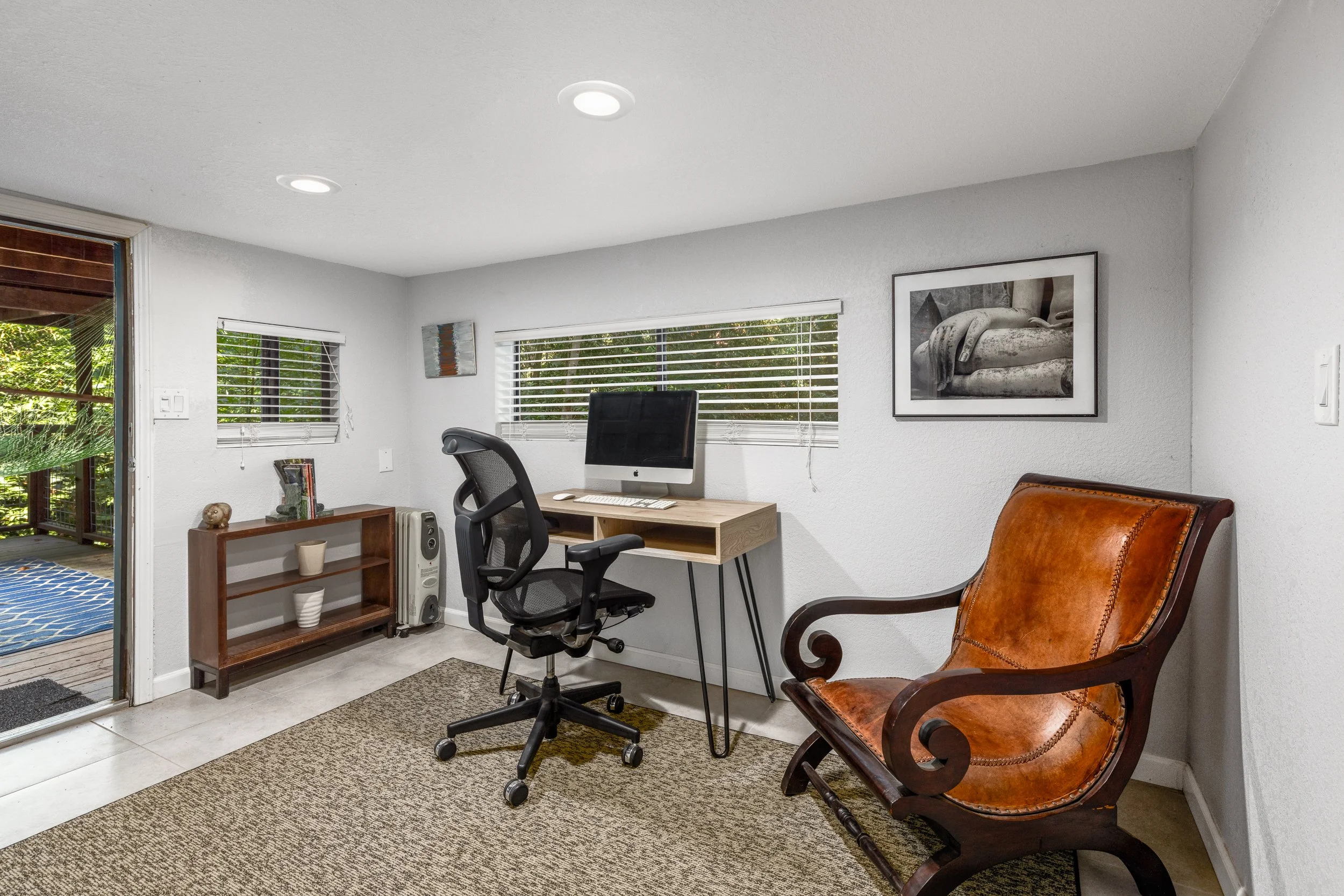 A home office with white walls and a large window with horizontal blinds. The room has a wooden desk with a computer, a black ergonomic chair, a small wooden shelf with decorative items, a framed black and white photograph on the wall, and a brown le
