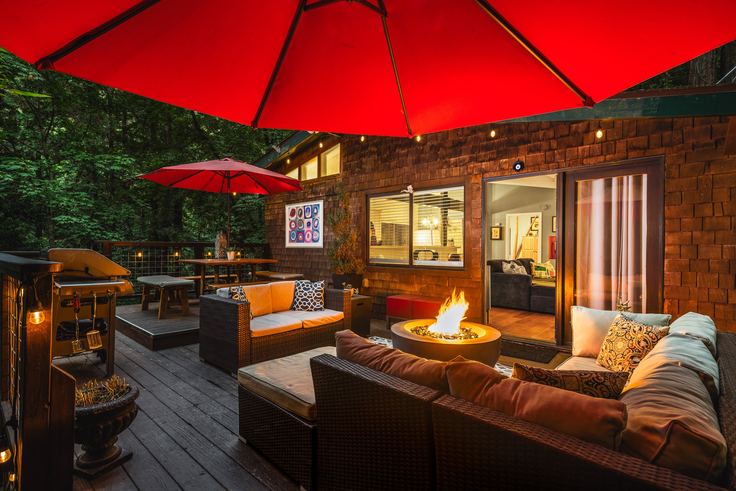 A cozy outdoor patio at night with red umbrellas, string lights, a firepit, and outdoor furniture, attached to a wooden house with large windows and colorful artwork inside.