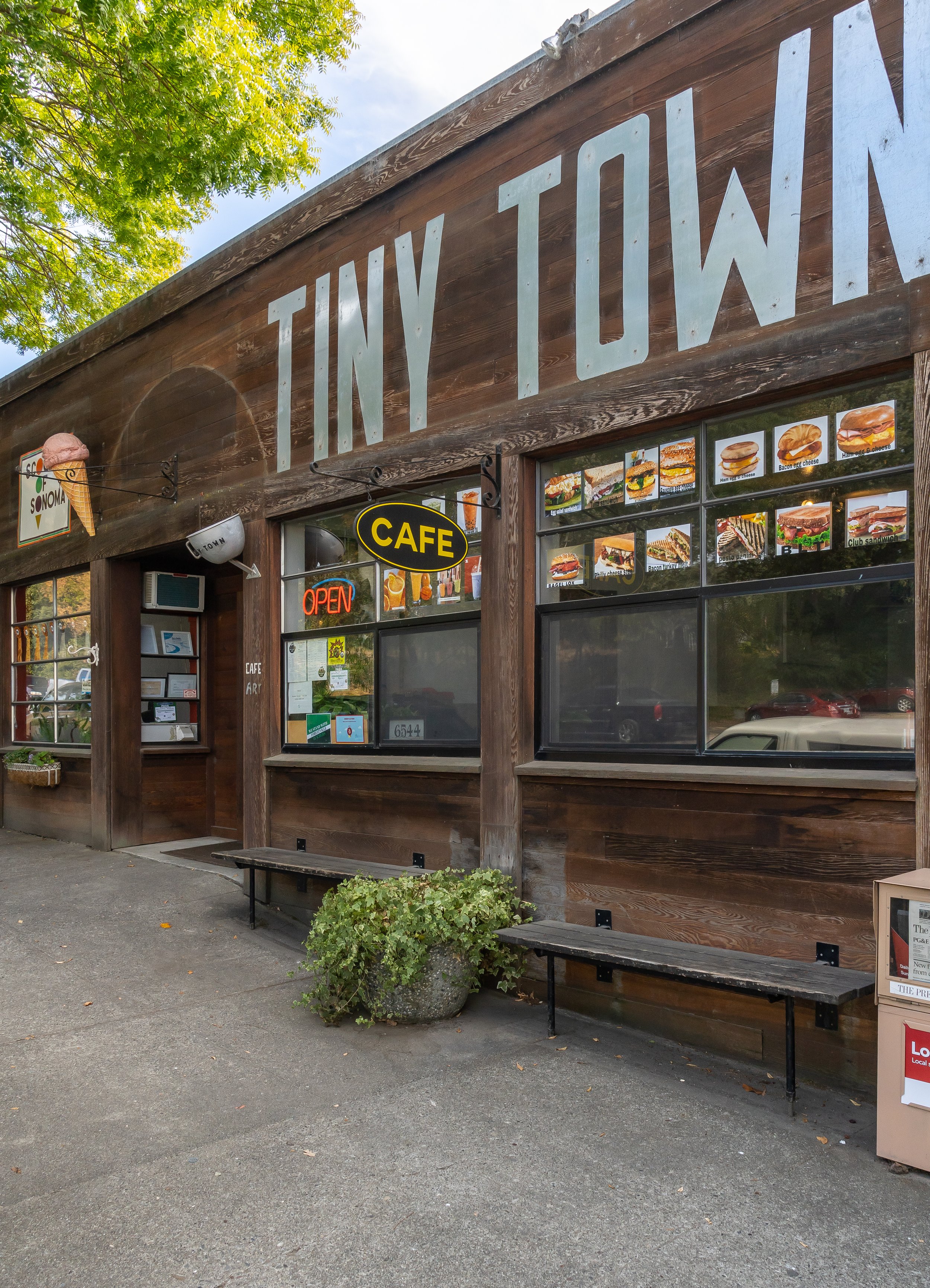 A wooden storefront with a large sign that reads "Tiny Town," featuring images of sandwiches, burgers, and ice cream. There is a neon "Open" sign in the window and a black sign with yellow text that says "Cafe." Outside, there are benches, a trash ca