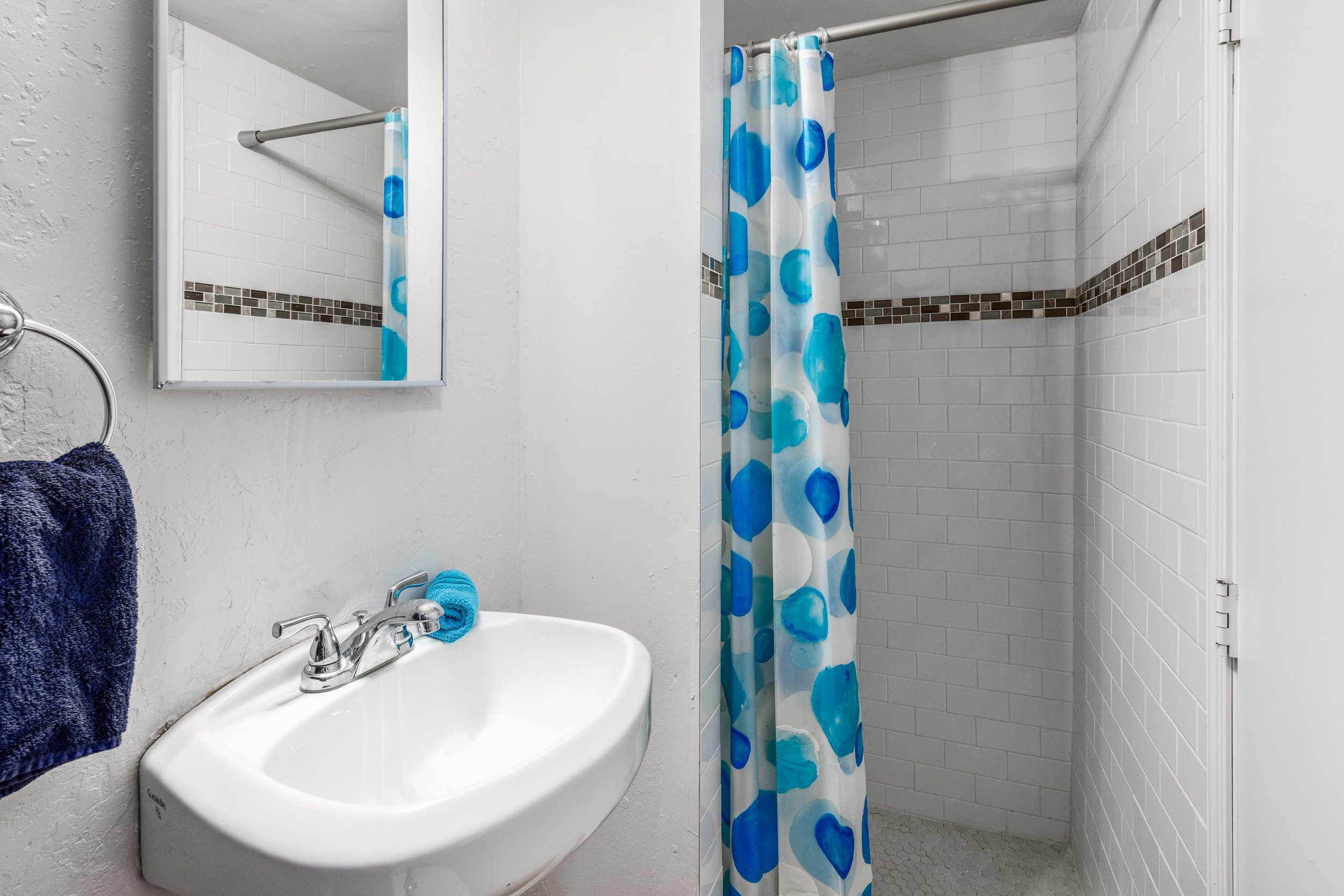 A small bathroom with a white sink, a mirror, a dark blue towel hanging on a towel ring, a blue washcloth on the sink, a shower with a colorful blue and white polka dot shower curtain, white tiled walls with a decorative dark tile border, and a small