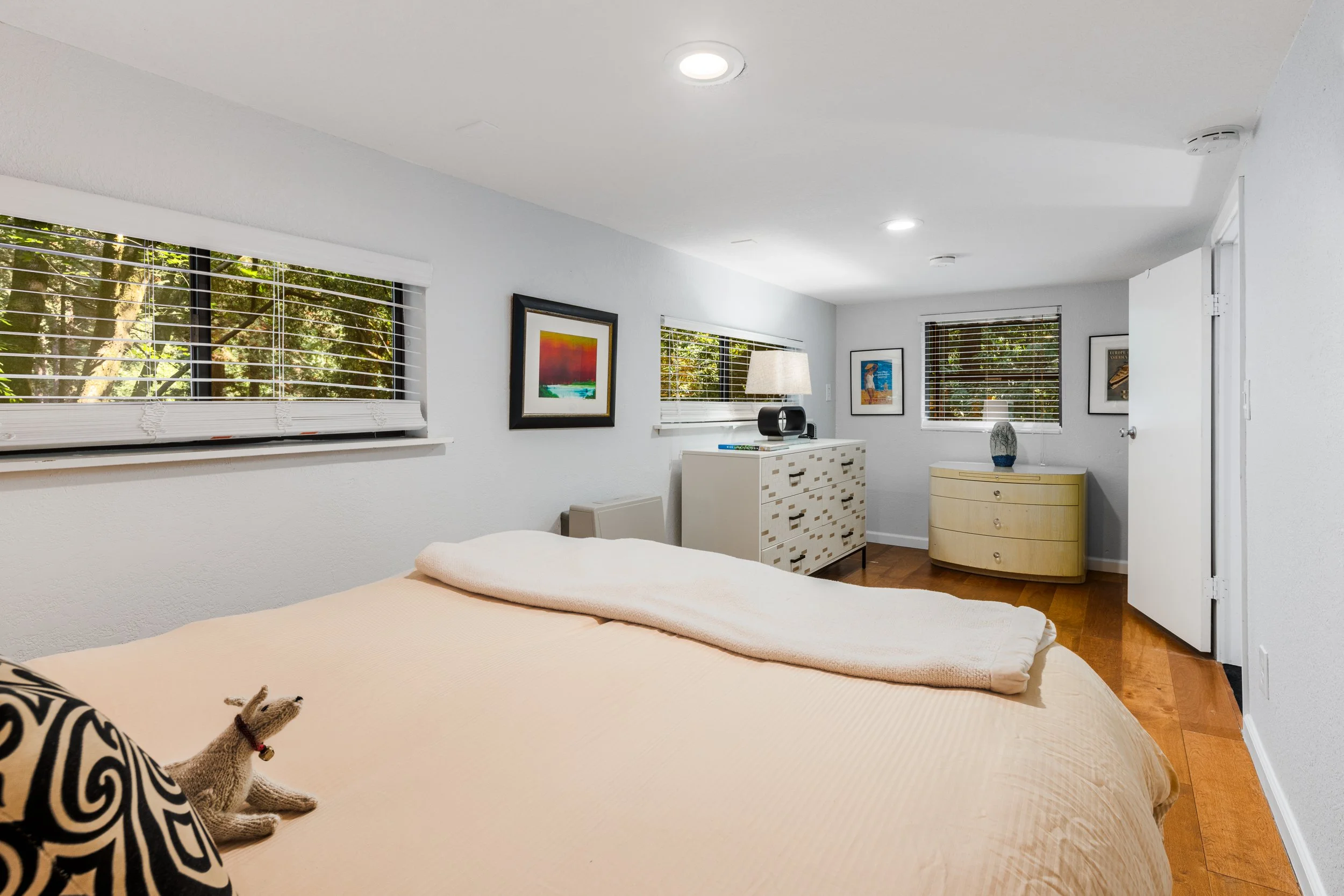 A bedroom with white walls and wooden flooring, featuring a bed with a beige bedspread and decorative pillows, including a patterned black and white pillow with a stuffed animal on it. There are three windows with white blinds letting in natural ligh