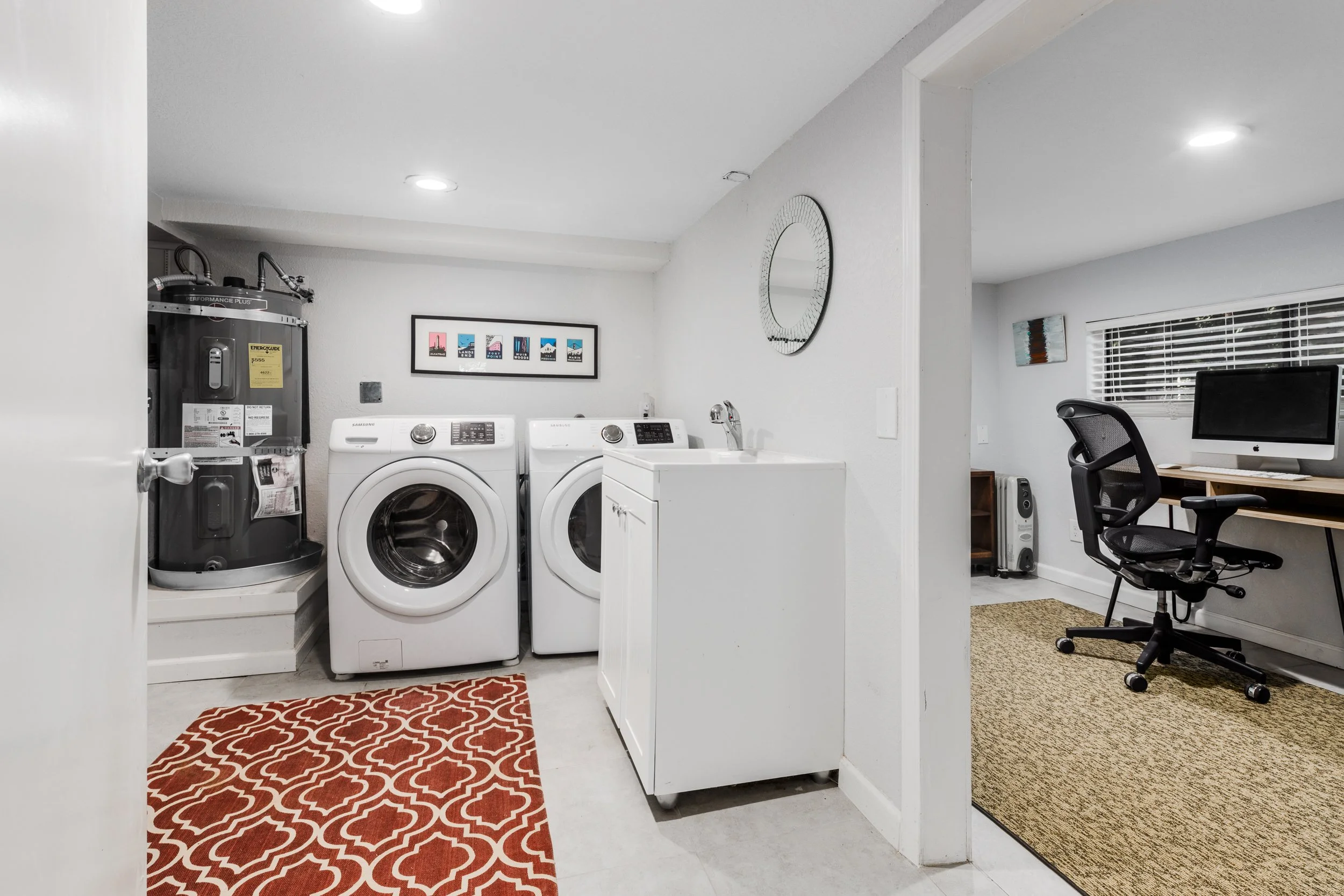 A laundry room with a white washing machine and dryer, a utility sink, a framed picture on the wall, a red patterned rug, and a workspace with a computer and chair.