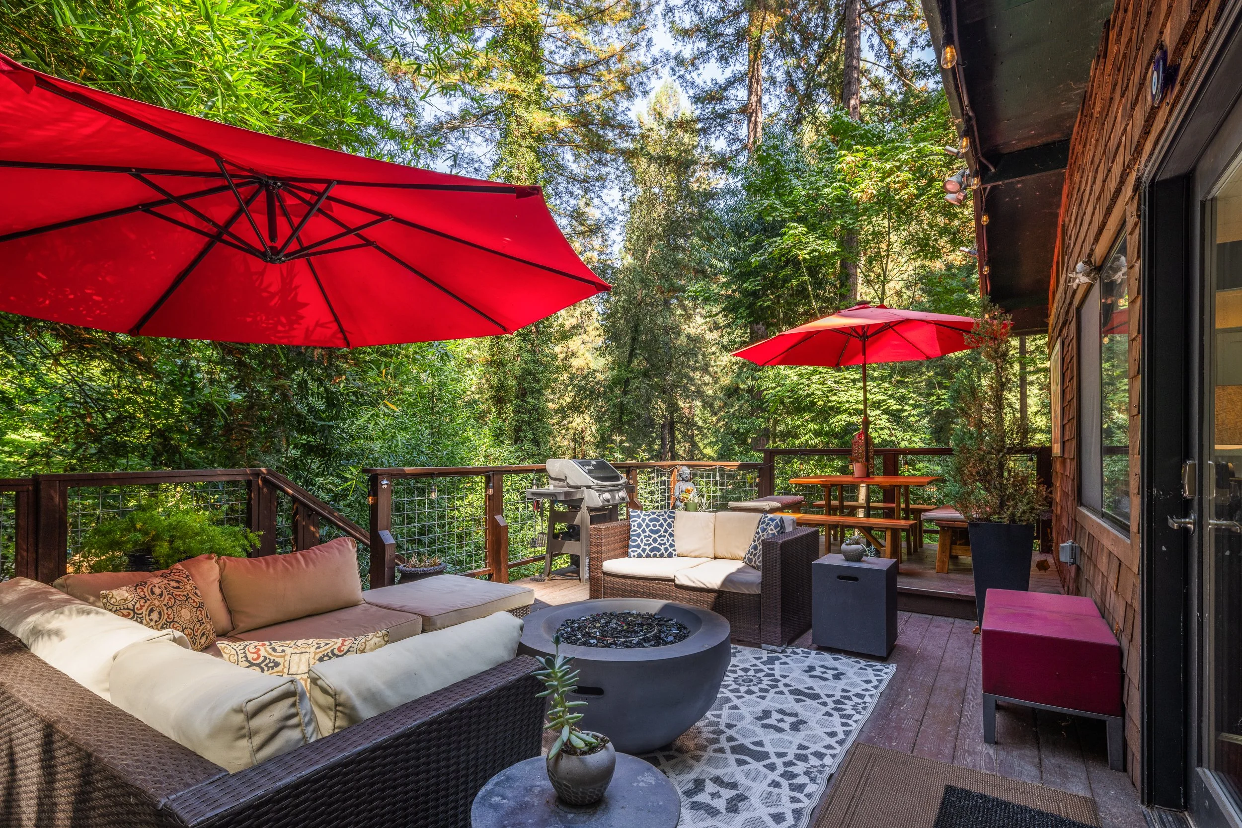 A cozy outdoor porch with outdoor furniture, red umbrellas, and surrounded by tall trees and greenery.