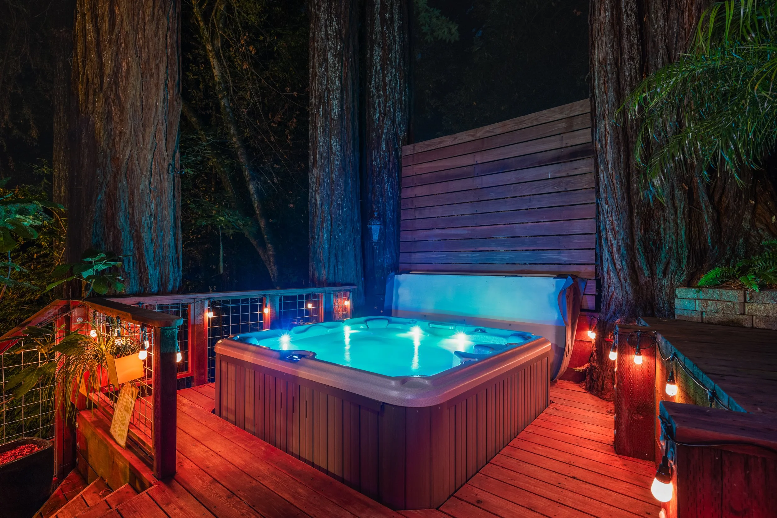 Outdoor hot tub in a wooden deck surrounded by large trees and string lights at night.