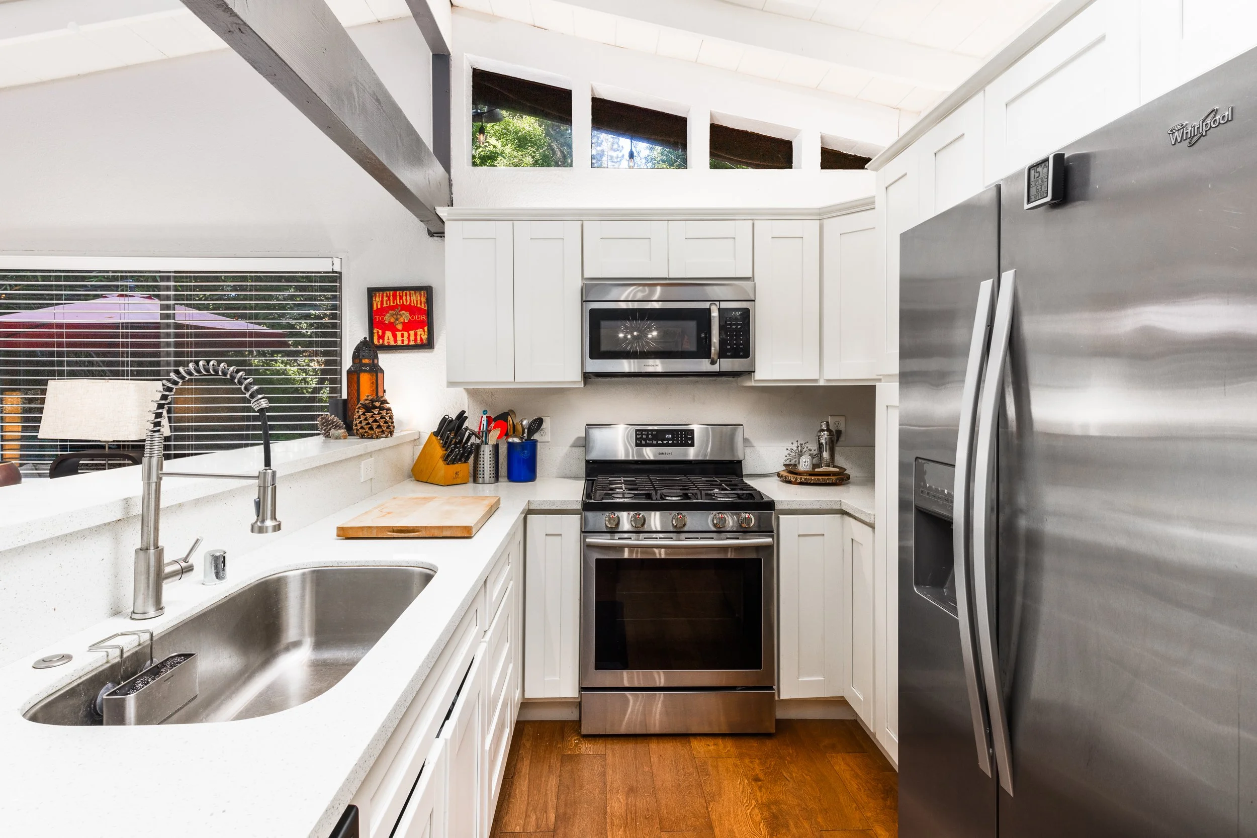 Modern kitchen with white cabinets, stainless steel appliances including microwave, stove, and refrigerator. Small window near ceiling, decorative items on counter, and wood flooring.
