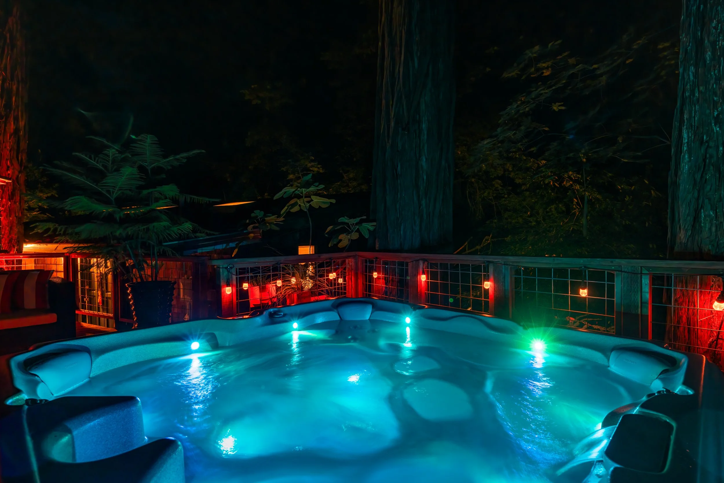 Nighttime scene of a backyard with a hot tub illuminated by blue lights, surrounded by a deck with string lights, a railing, and tropical plants, overlooking a dense forest with tall trees.