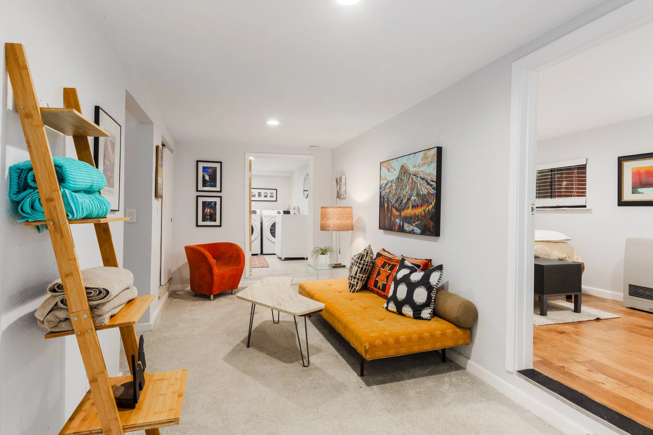 Interior view of a living room with a yellow mid-century modern sofa, a glass coffee table, and colorful patterned pillows. A red velvet armchair, a tall floor lamp with a beige shade, and wall art including a landscape painting are visible. The room