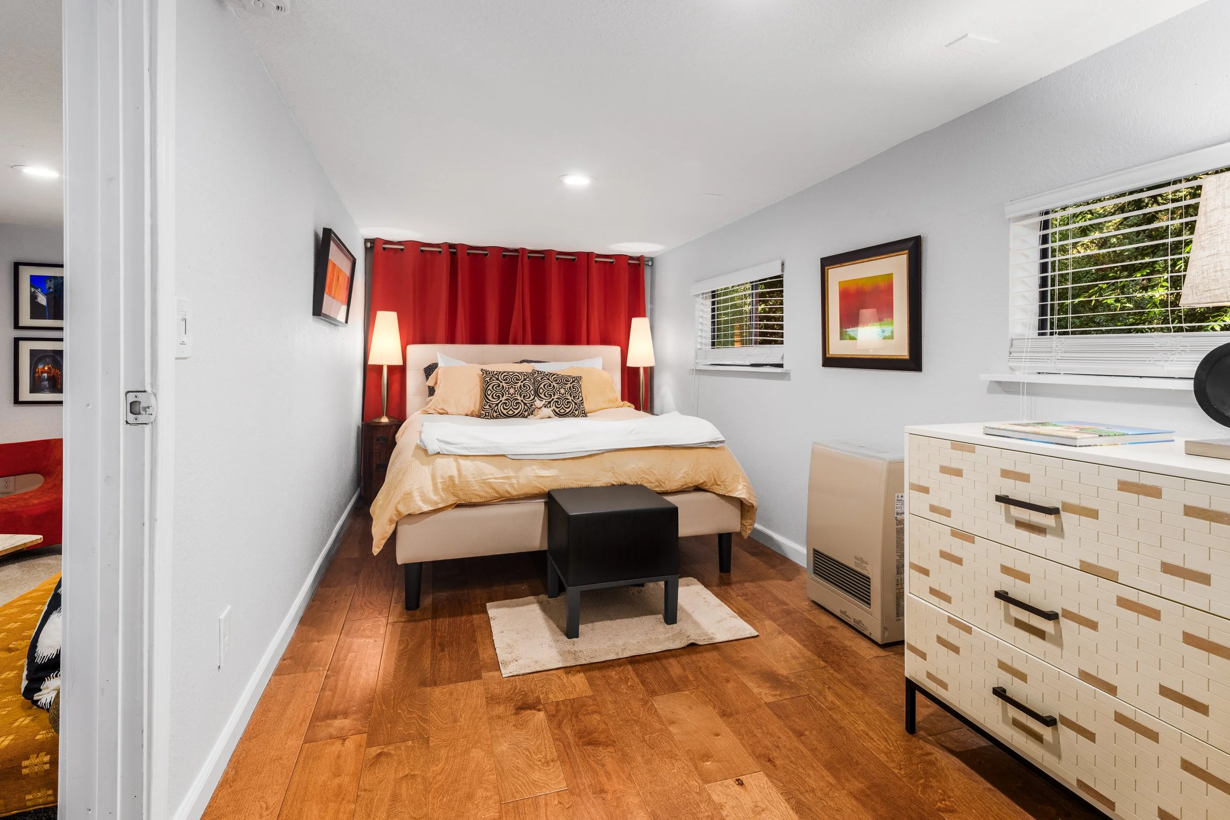 A bedroom with a bed featuring a beige headboard, orange bedding, and decorative pillows. There are two tall lamps on either side of the bed, and the room has white walls, wooden flooring, and two windows with blinds and a picture frame on the wall. 