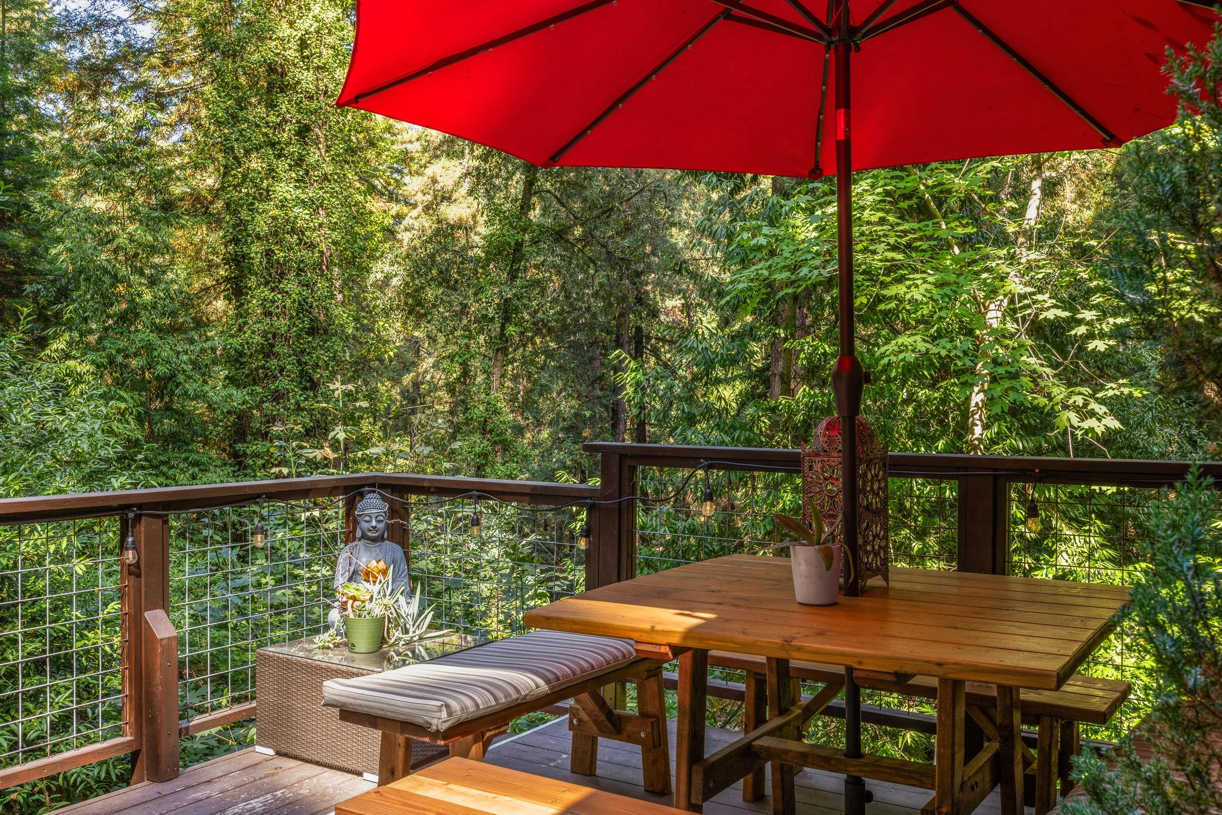 A wooden deck with outdoor furniture including a table, benches, and a potted plant under a large red patio umbrella, surrounded by lush green trees.