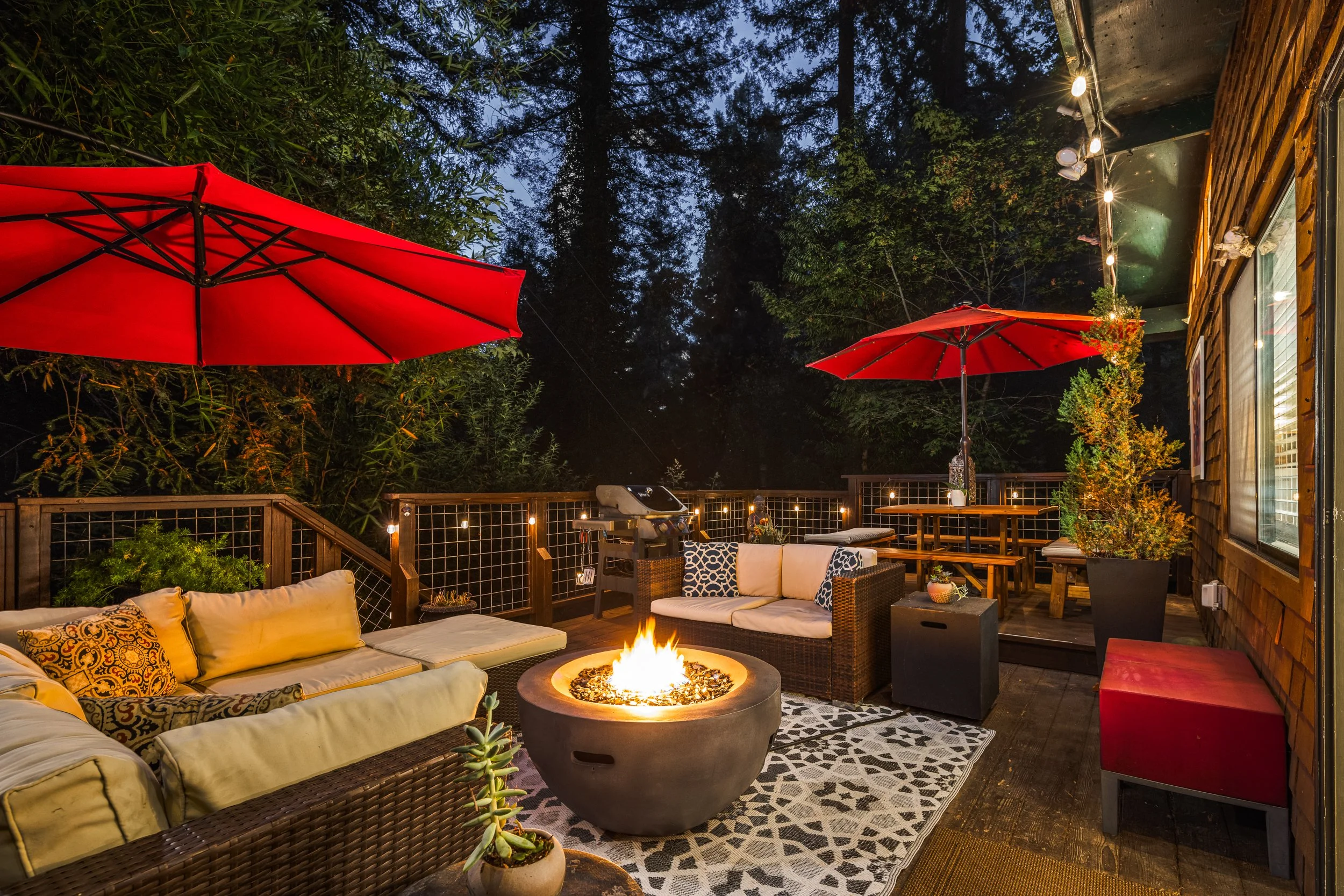 A cozy outdoor patio at night with a firepit, beige cushioned outdoor sofa, wicker armchair, red umbrellas, string lights, and lush greenery surrounding the space.