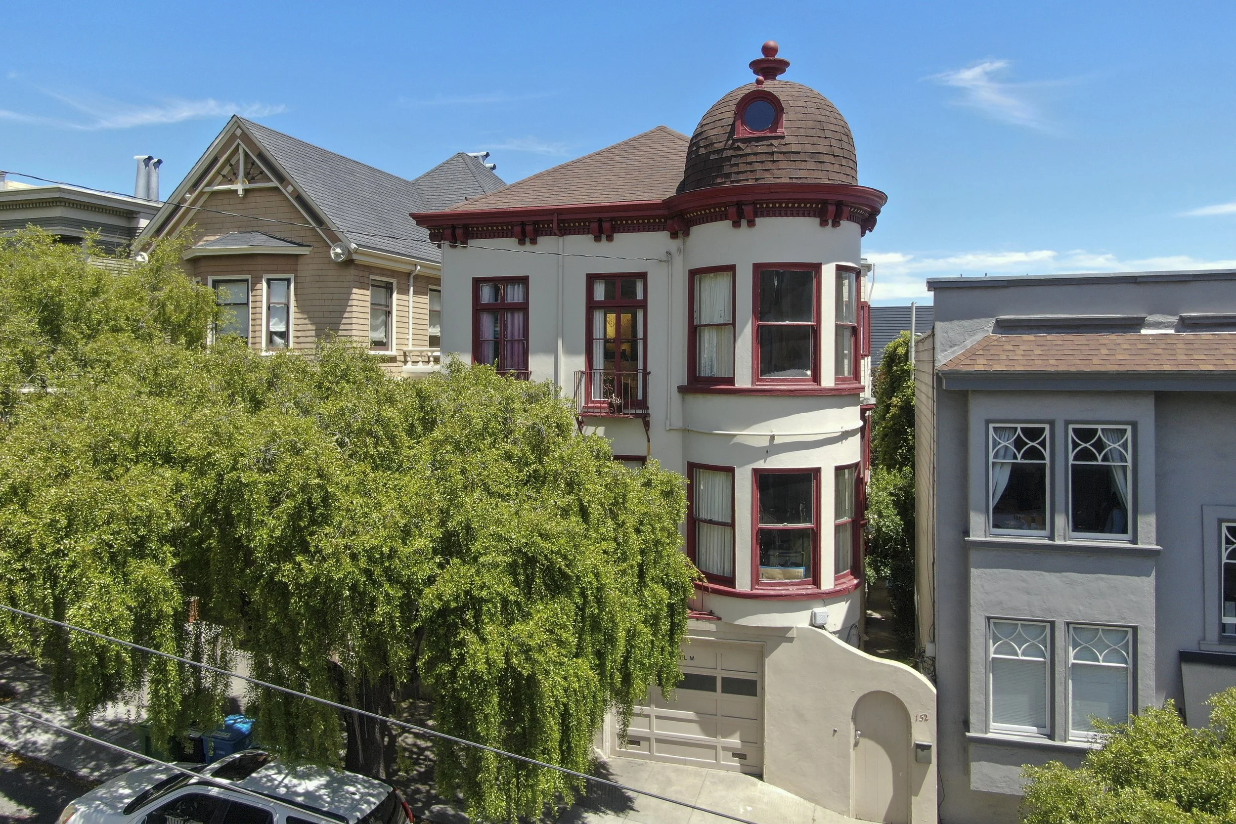 Colorful Victorian-style house with rounded turret and red accents, surrounded by trees and neighboring houses on a sunny day.