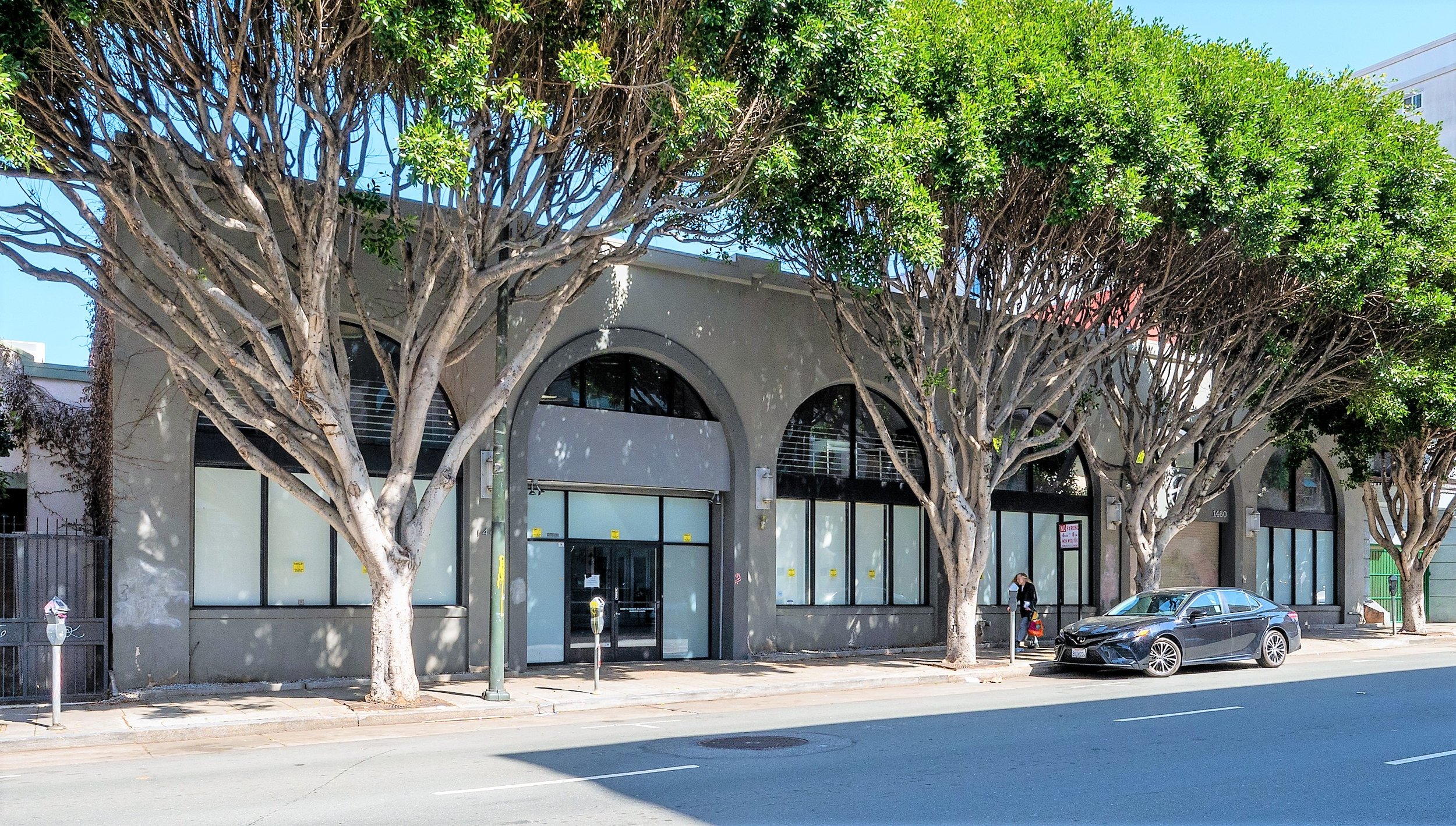 Street view of a commercial building with large arched windows, surrounded by tall leafy trees, a parked black car, and a woman walking on the sidewalk.