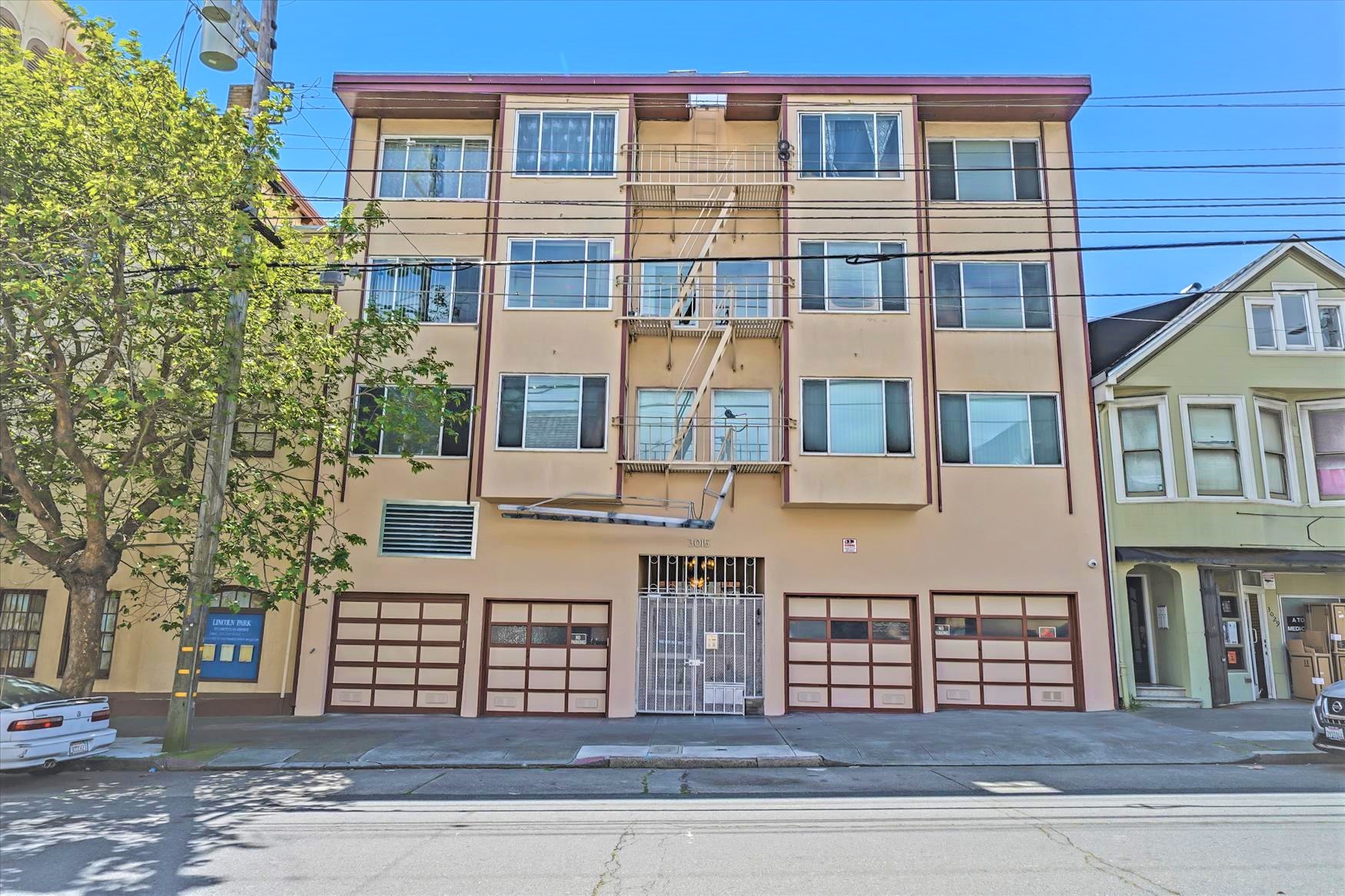 multi-story beige apartment building with multiple windows, fire escapes, and garage doors at street level, next to a green house and a tree on a sunny day.