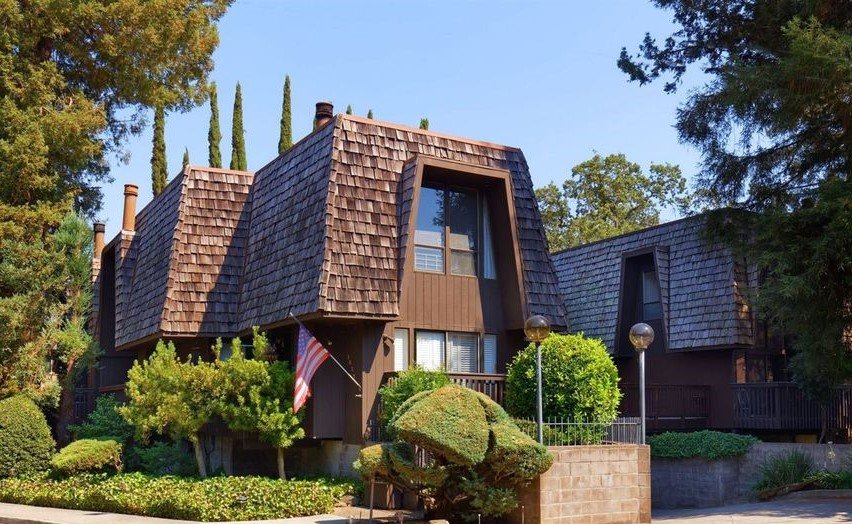 A brown A-frame house with a steep roof and shingle siding, surrounded by lush green bushes and trees, with two lampposts in front.