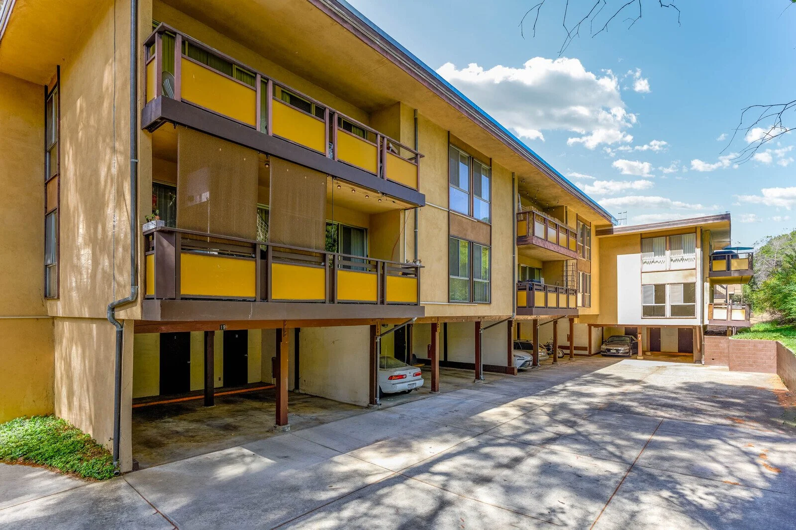 Apartment complex with yellow walls, balconies, and parking area with cars underneath, surrounded by trees and clear sky.