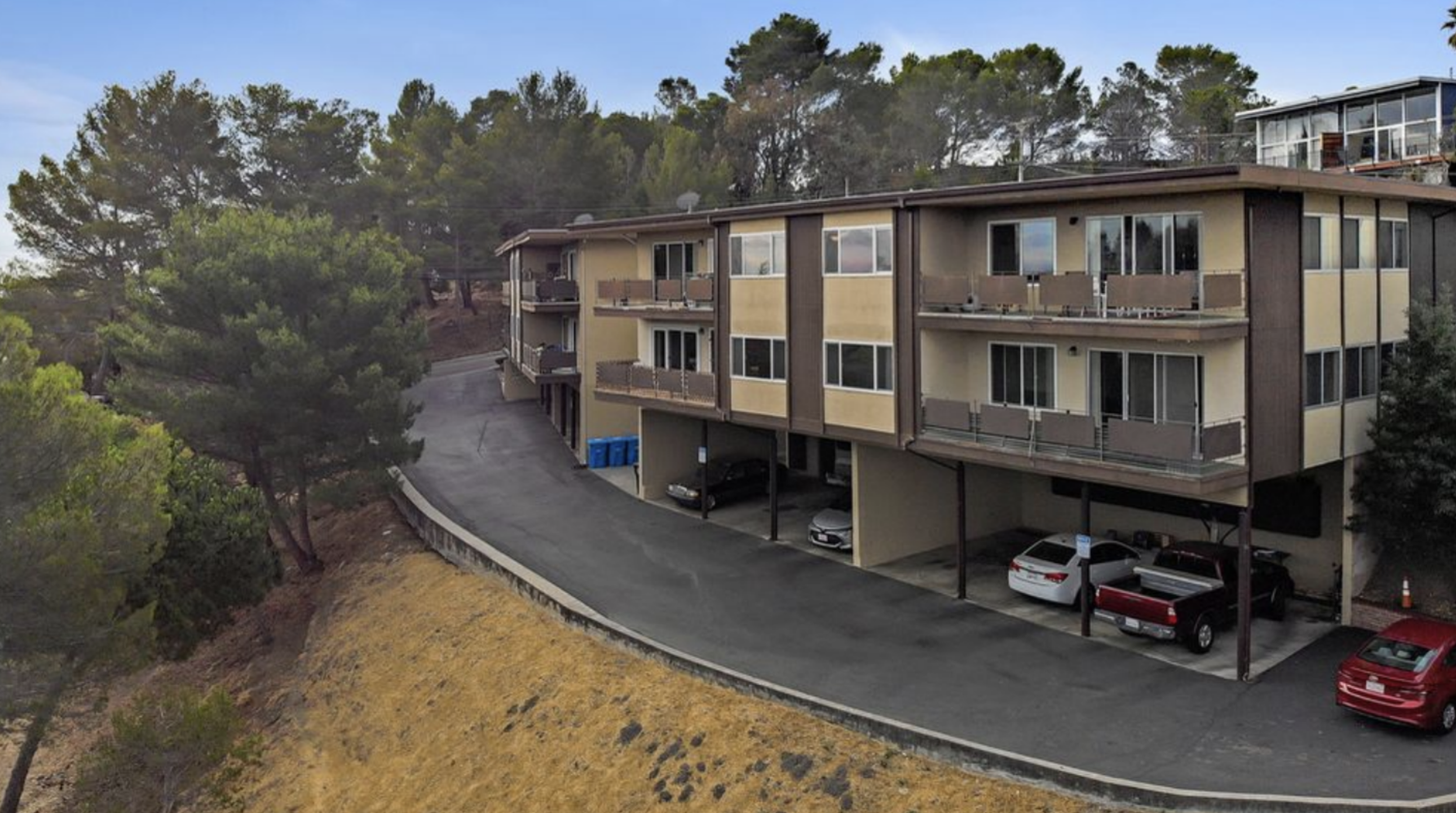 A multi-story residential building with parking underneath, parked cars, surrounded by trees and a hillside, with a clear sky.