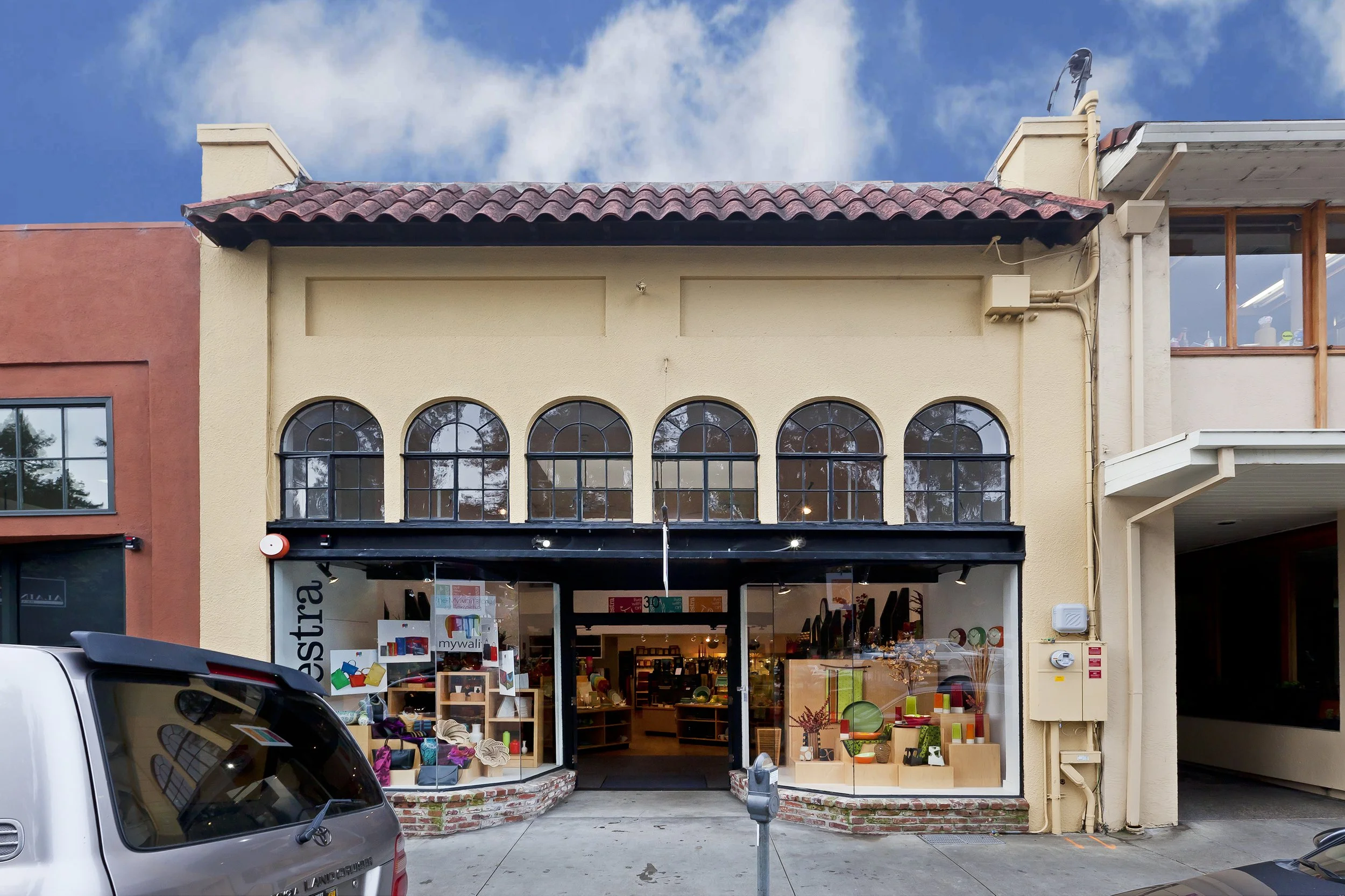 Street view of a storefront with large display windows and arched upper windows, with cars parked in front and a partly cloudy sky above.