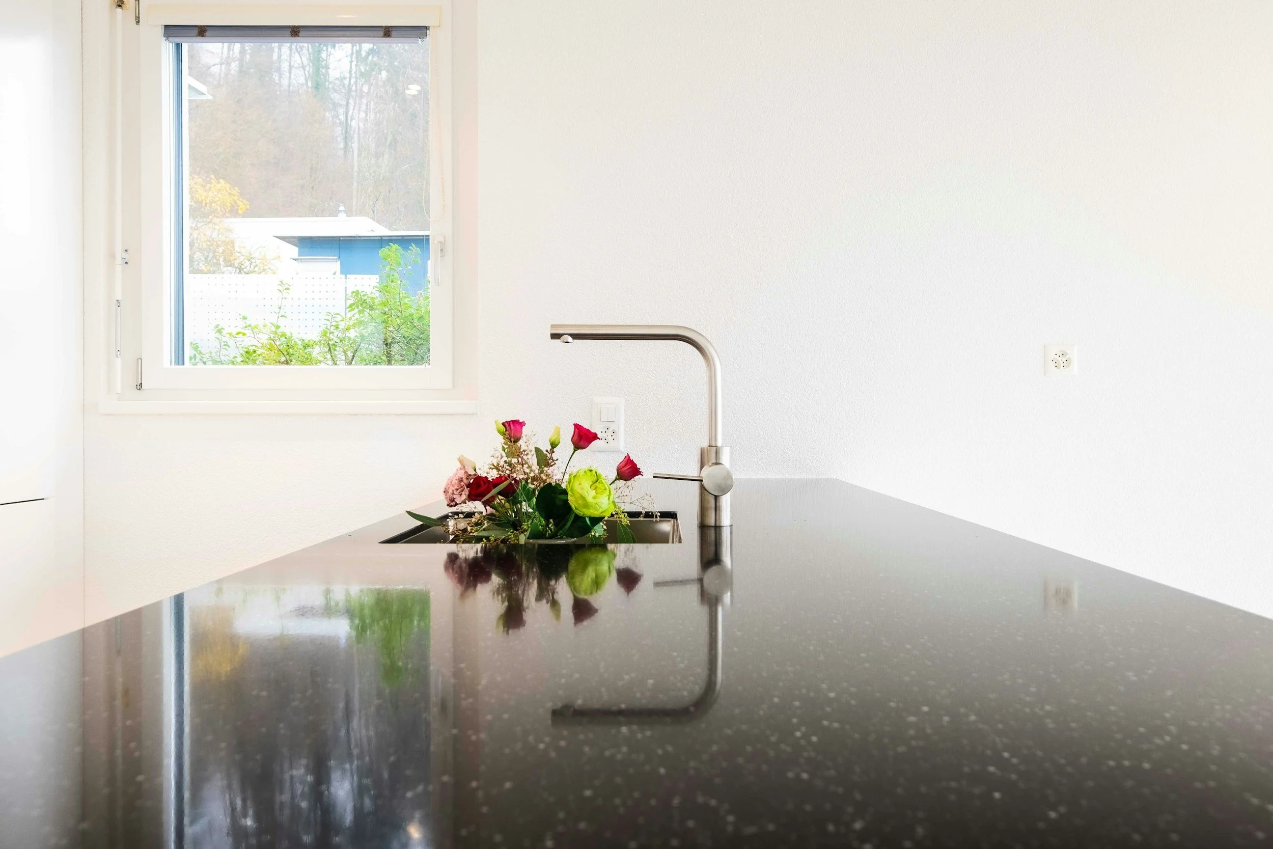 A modern kitchen with a black speckled countertop, a metallic faucet, and a bouquet of pink, red, and green flowers, with a window showing trees and a blue building outside.