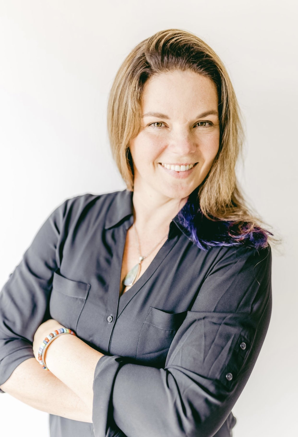 A woman with shoulder-length light brown hair and light skin, smiling, wearing a black button-up shirt, with her arms crossed and several colorful bracelets on her left wrist, standing against a plain white background.