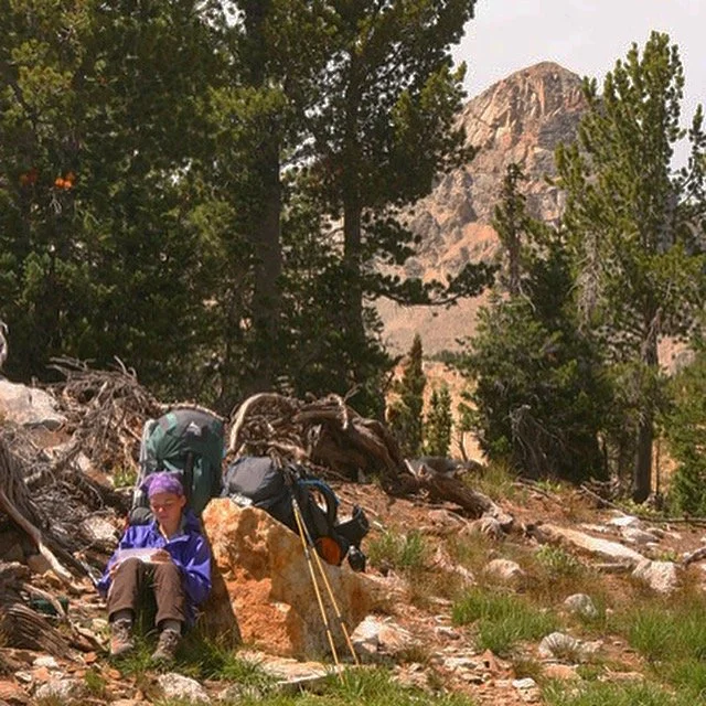 A woman pausing to write in her journal with a purple bandana and green backpack sitting on the ground, surrounded by trees and mountain scenery.