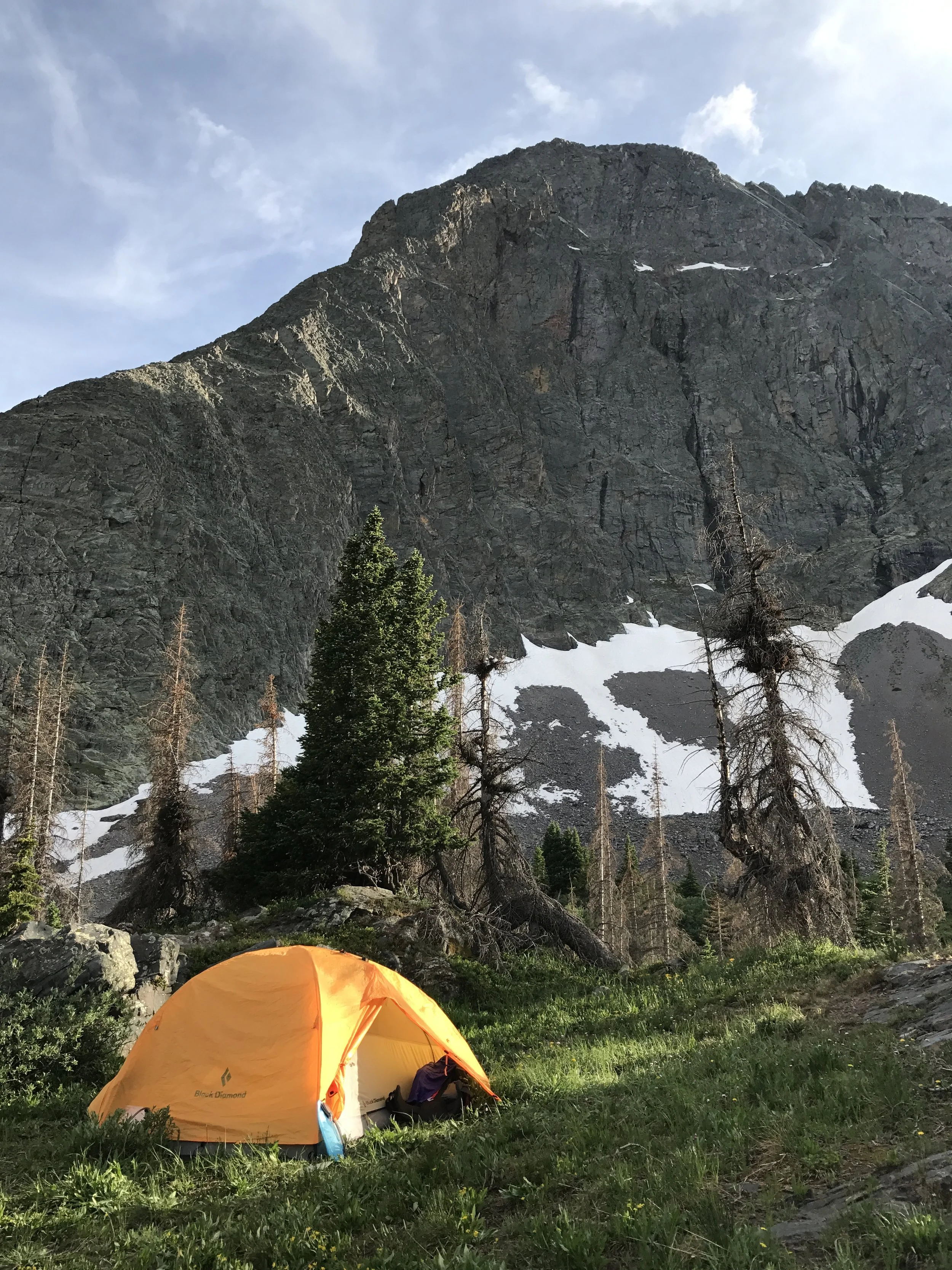 An orange camping tent set up on green grass near a mountain with patches of snow and trees, under a partly cloudy sky.