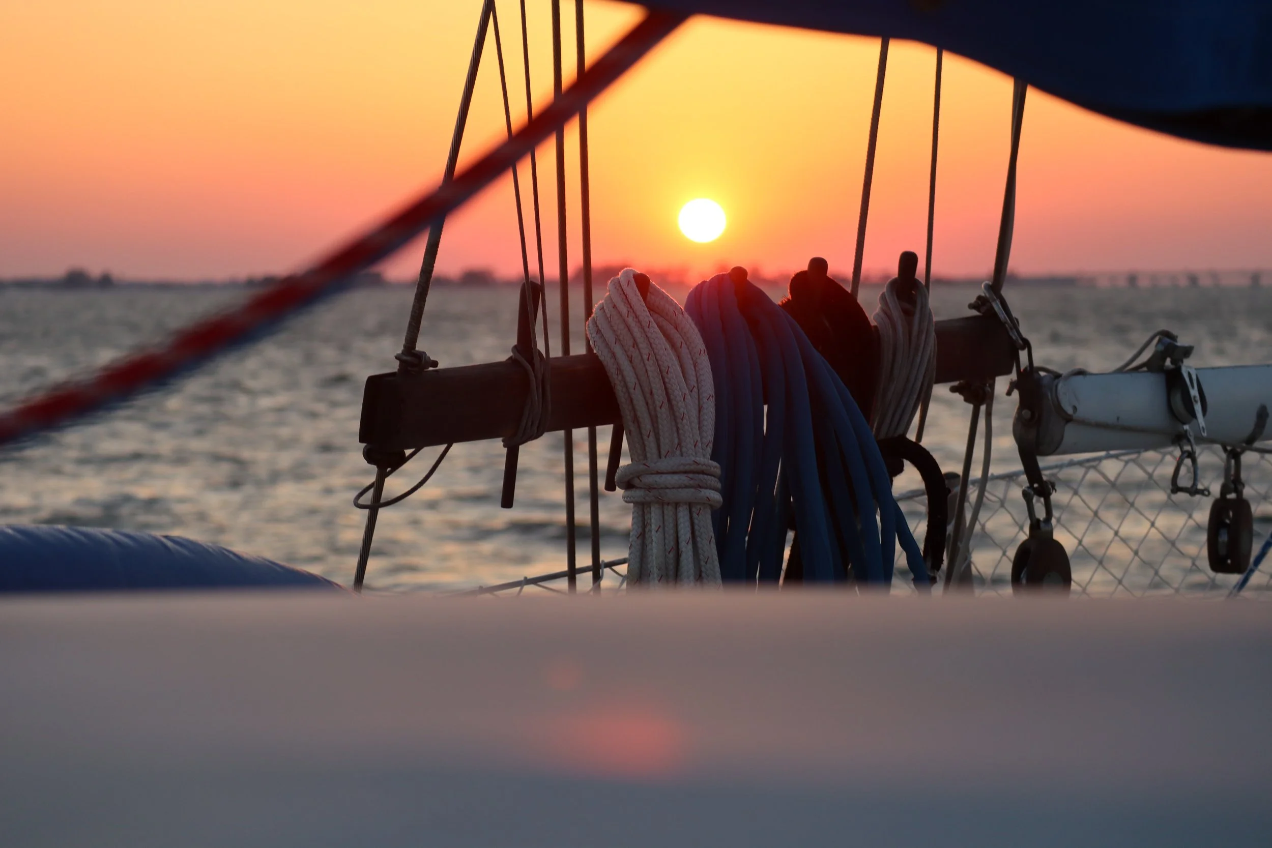 Close-up of ropes and rigging on a sailboat with a sunset over water in the background.
