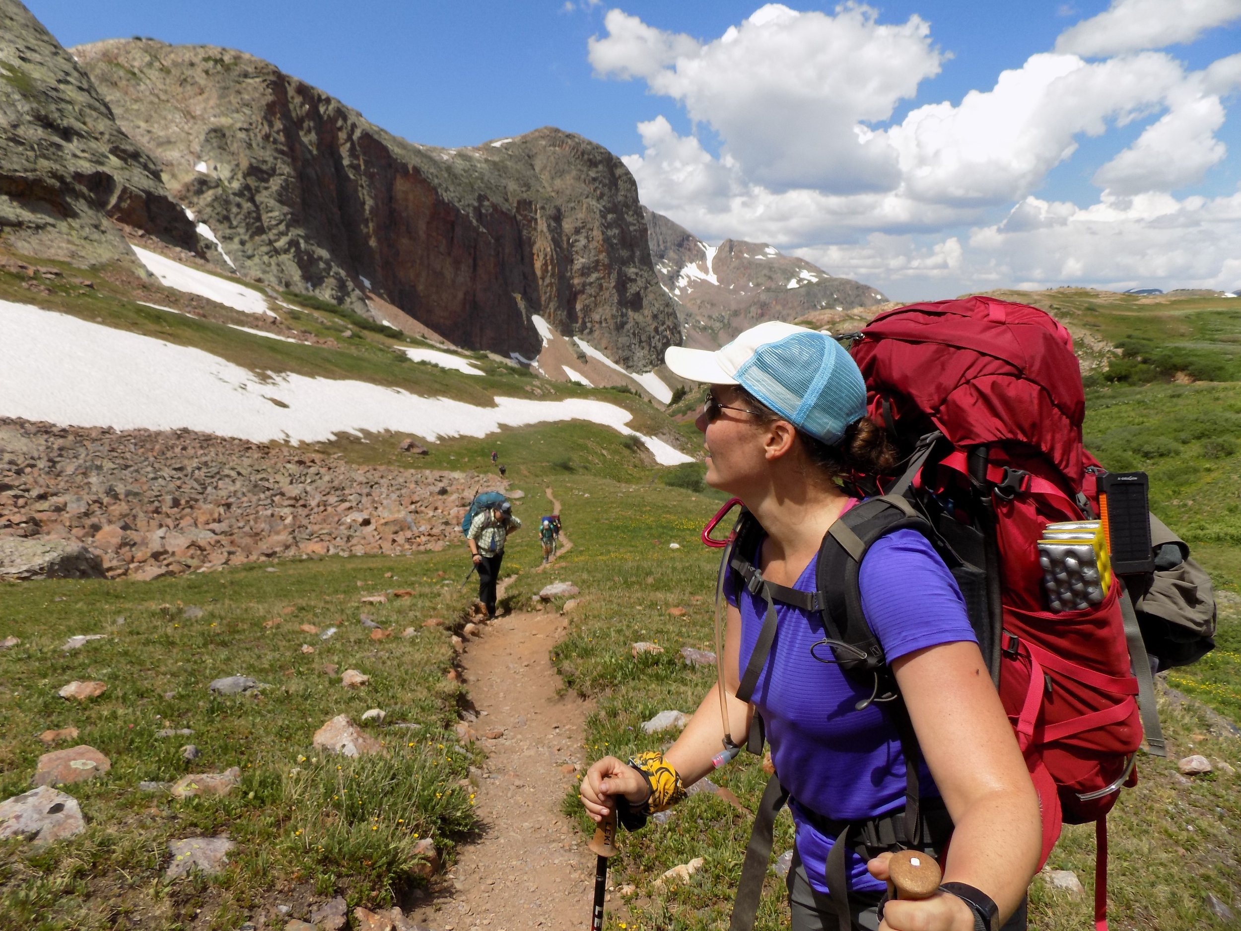 A woman hiking in a mountainous area with snow patches, wearing a blue cap, sunglasses, a purple shirt, and a red backpack.