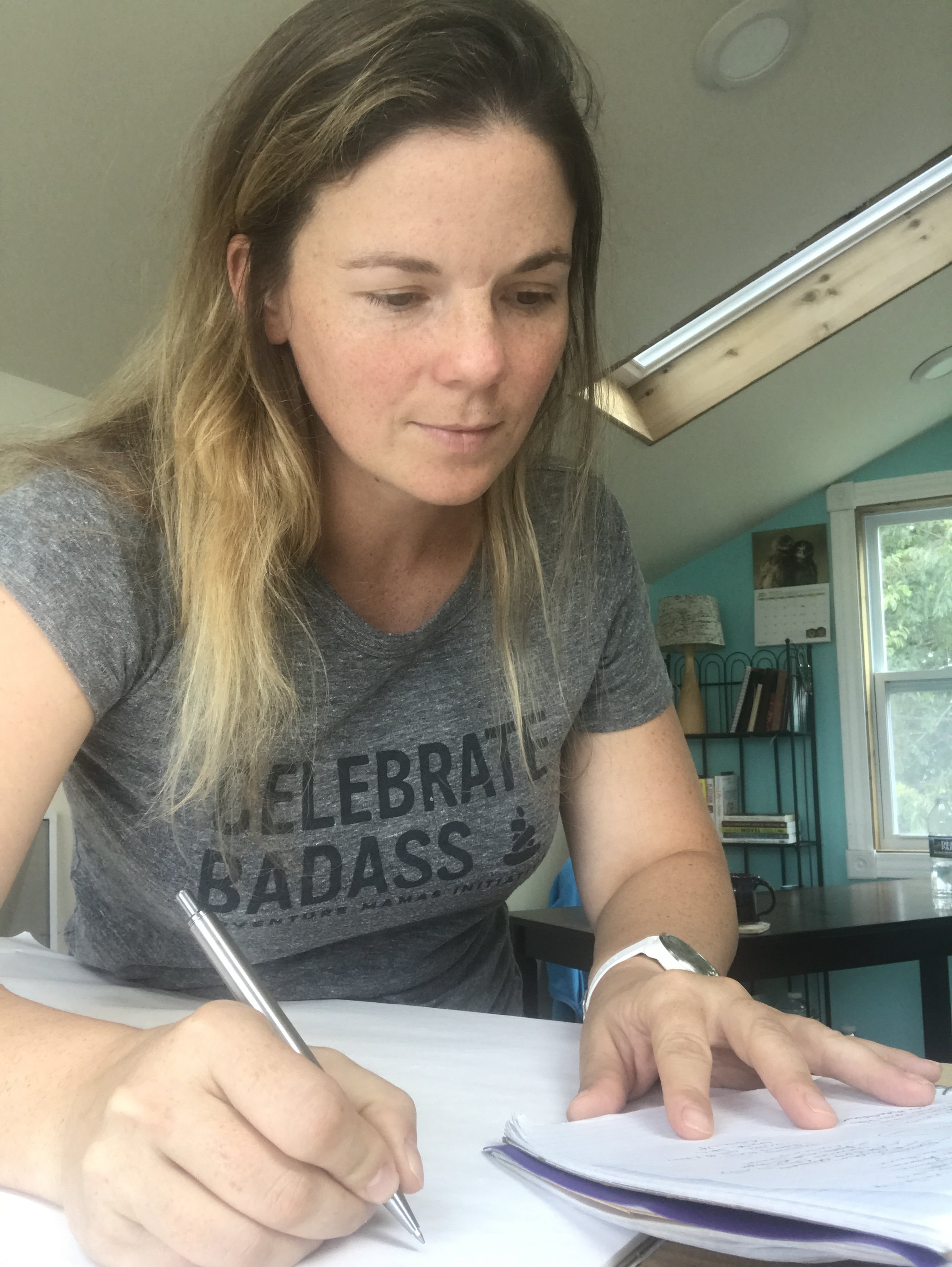 Close-up of woman writing on paper with a pen, wearing a t-shirt with text saying "Celebrate Badass," seated at a desk in a room with a window and bookshelf in the background.