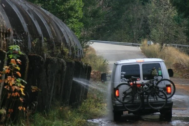 A white van parked on a muddy roadside with a bicycle mounted on the back, near a huge water pipe that's sprung a leak. The van gets a quick wash from the leak.