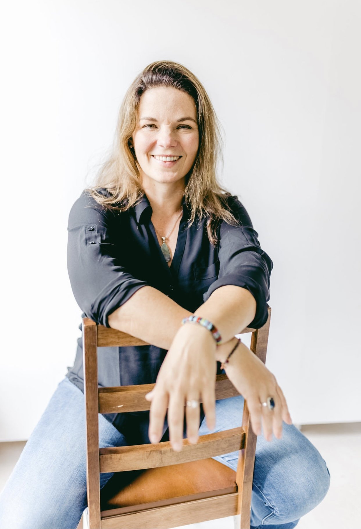 Woman with long, wavy light brown hair smiling, sitting on a wooden chair, in a black shirt and blue jeans, against a plain white background.