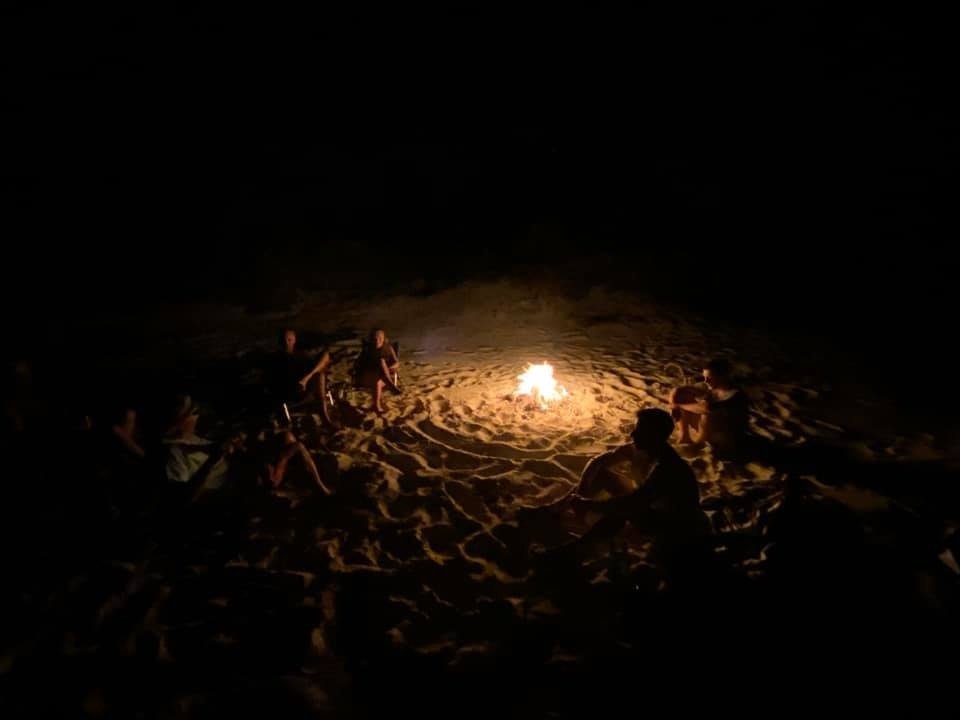 People sitting around a campfire on sandy ground at night, with only the fire illuminating the scene.
