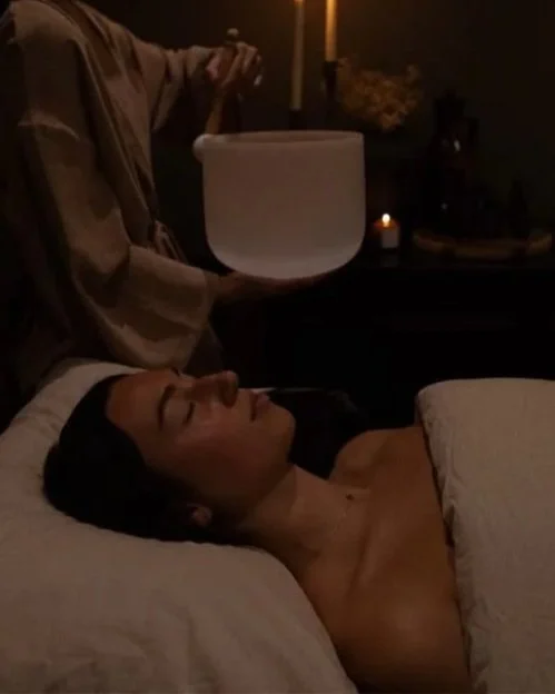 A woman lying in bed with eyes closed, having a sound healing session with a person holding a singing bowl.