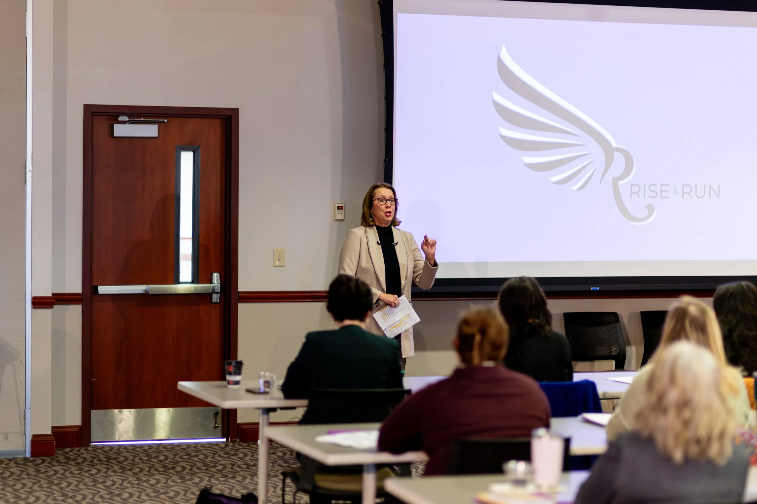 A woman standing and speaking in front of an audience in a conference room. She is holding papers and gesturing with her right hand, while a large screen behind her displays a logo with the words 'RISE & RUN' and a stylized wing design.