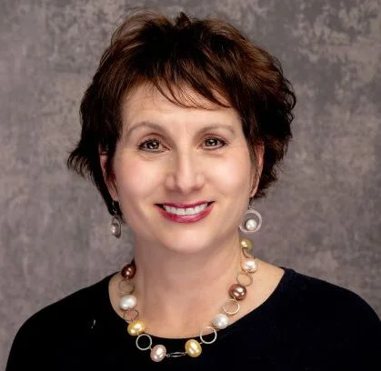 A woman with short, brown hair smiling, wearing pearl and gold jewelry, against a gray background.