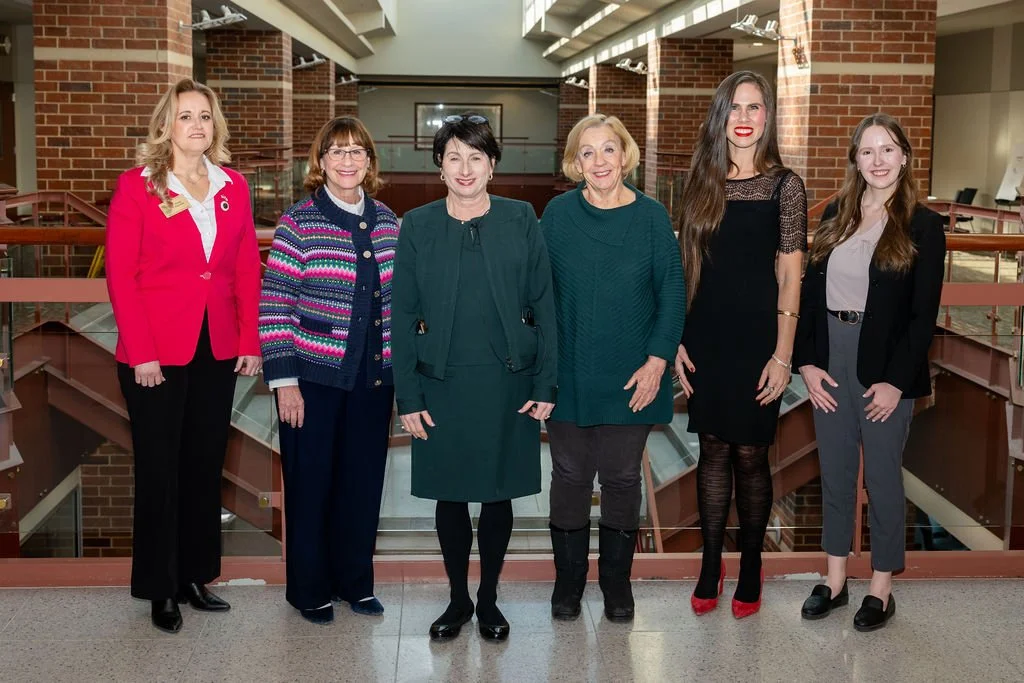 Six women standing side by side inside a building with brick walls and glass railing, posing for a group photo.