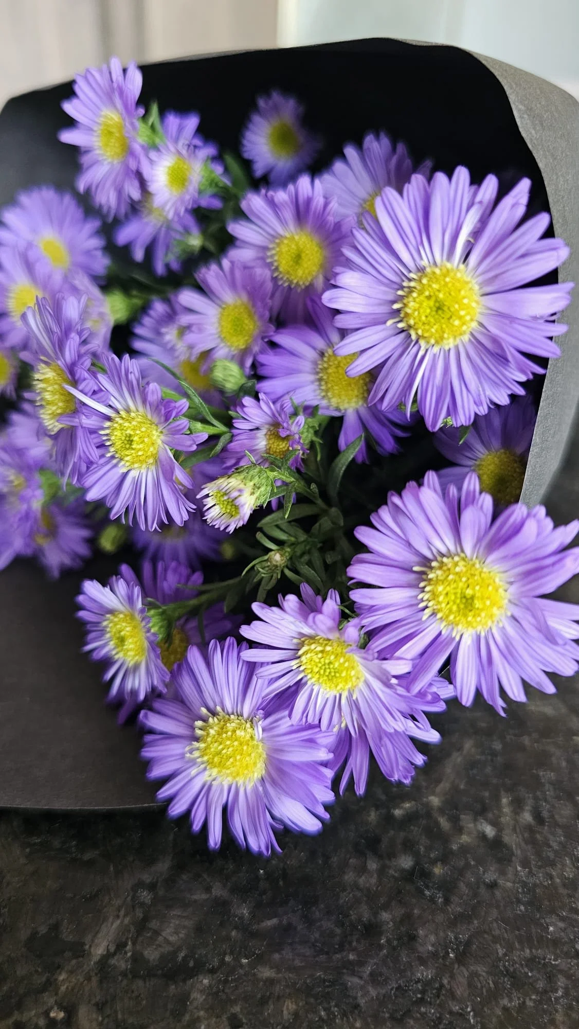 Un bouquet de fleurs violettes et jaunes, enveloppé dans du papier noir, posé sur une surface sombre.