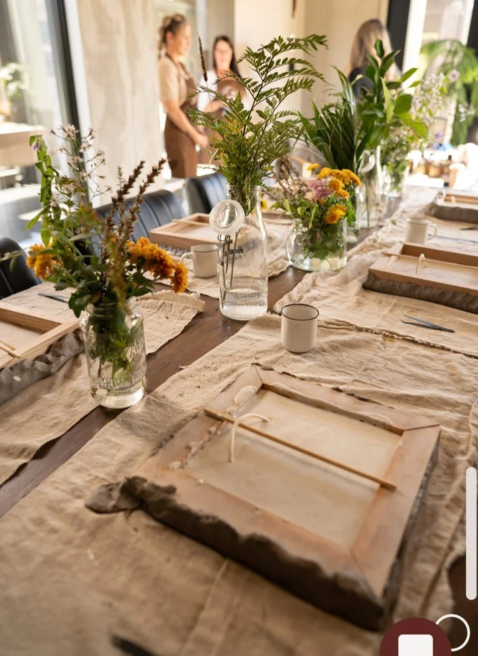 Une table en bois décorée avec des vases remplis de fleurs sauvages colorées, entourée de couverts et de tasses en céramique, dans une pièce lumineuse avec des personnes en arrière-plan